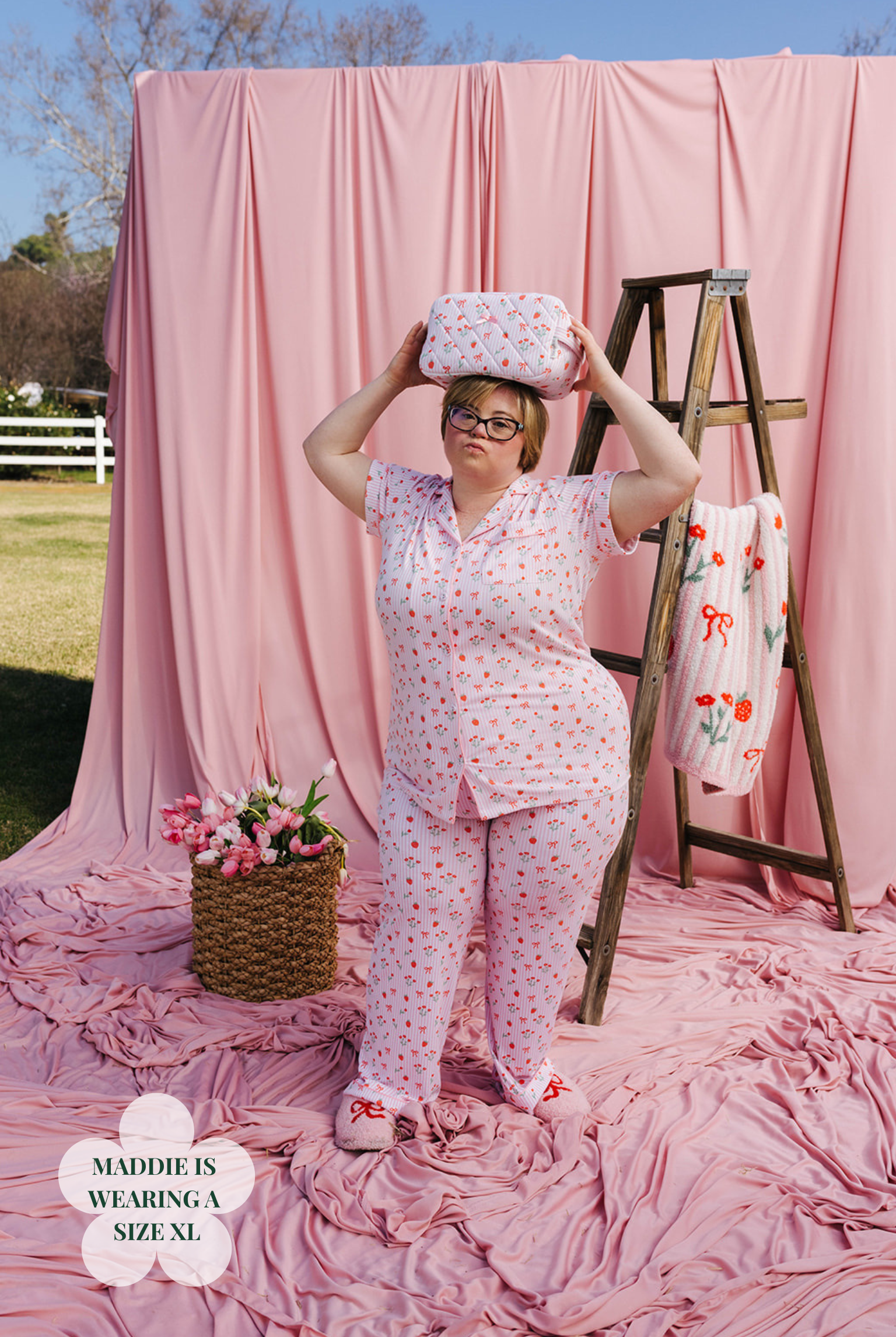 Woman in a pink floral outfit holding a box against a pink backdrop with a ladder and flowers.