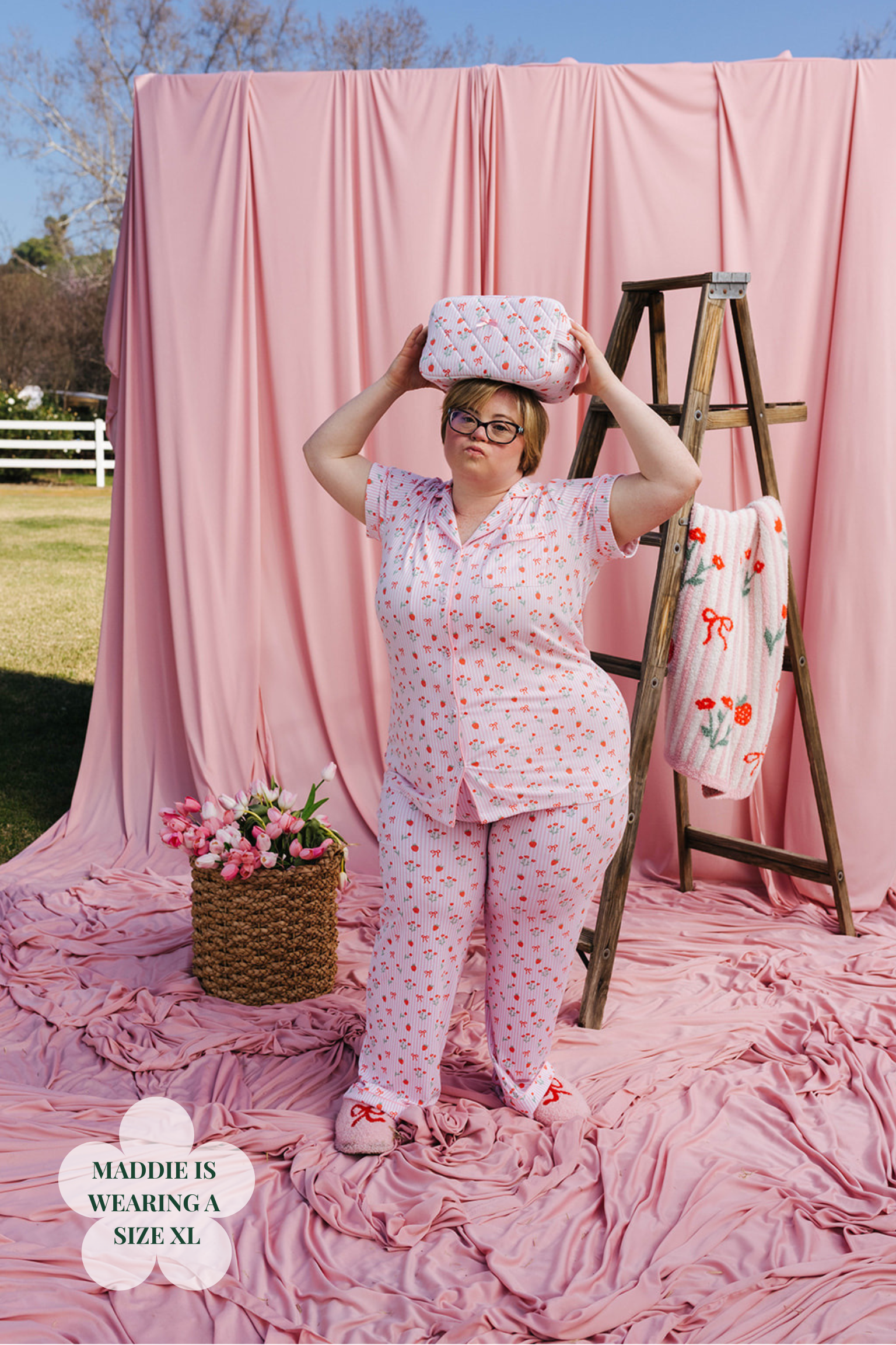 Woman in a pink floral outfit holding a box against a pink backdrop with a ladder and flowers.