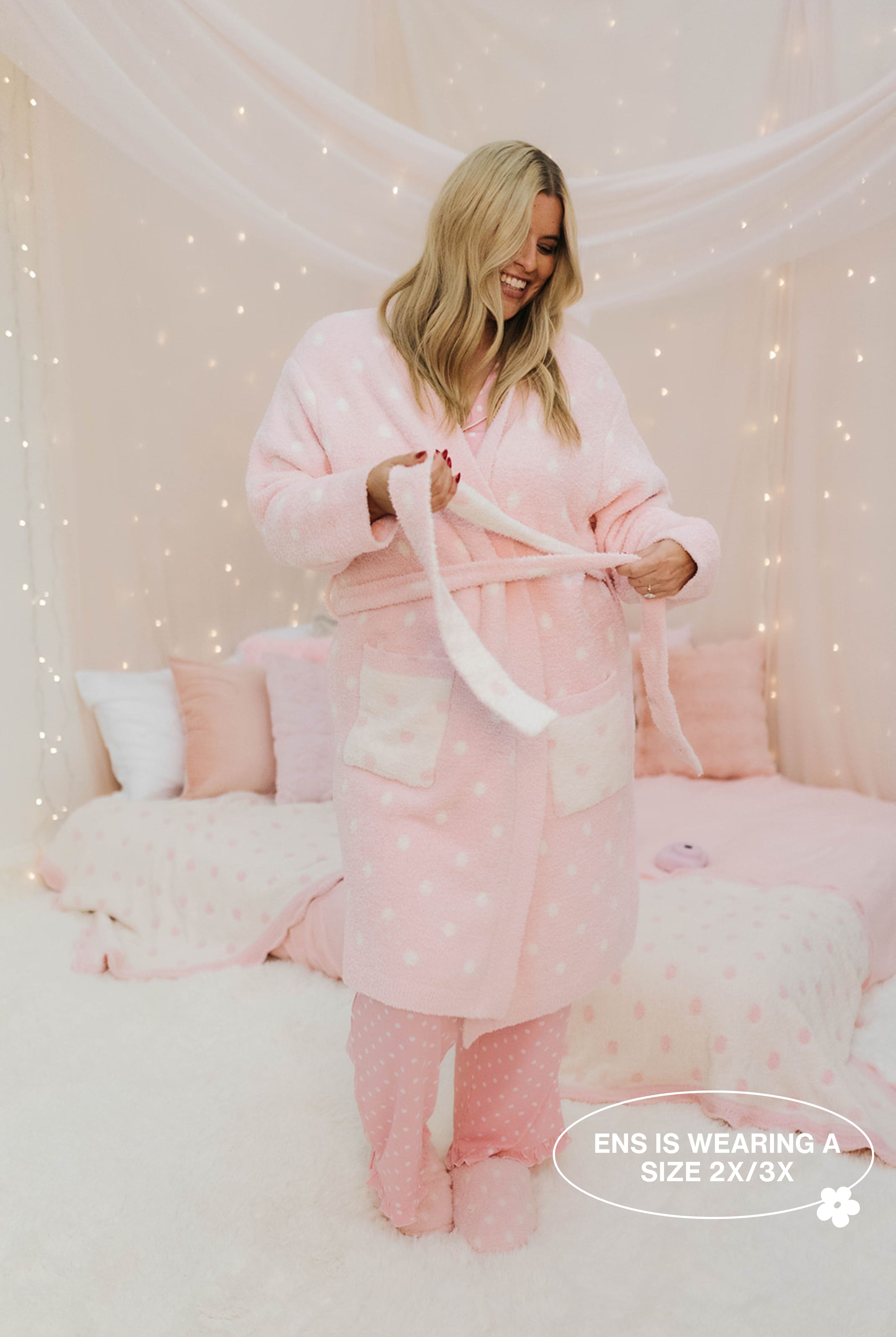 Woman wearing a pink robe with white polka dots in a cozy room with fairy lights.