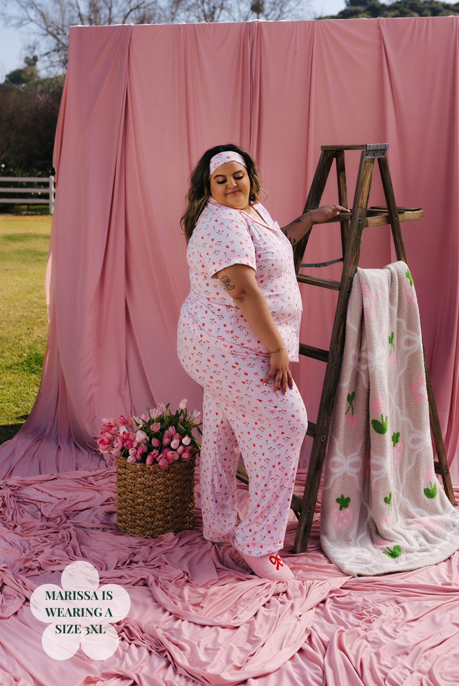Woman in a floral outfit standing next to a wooden ladder and pink fabric backdrop.
