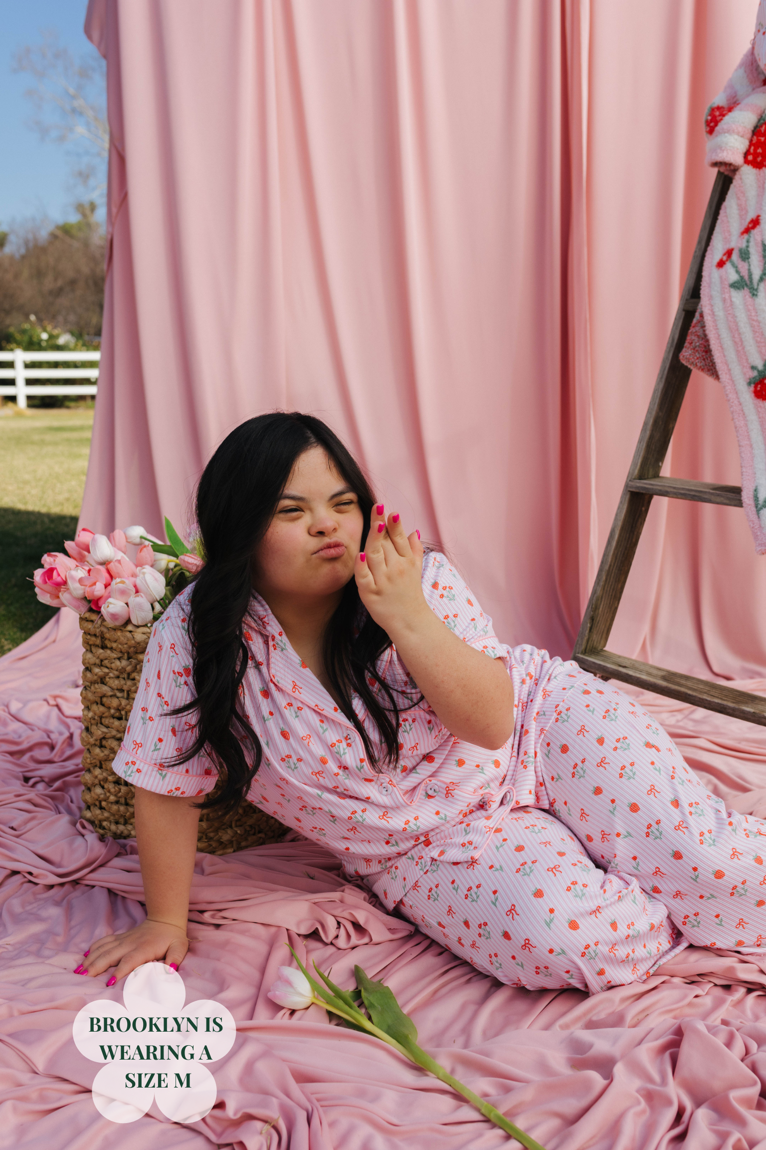 Woman in a floral pajamas sitting on a pink blanket with flowers and a ladder in the background.