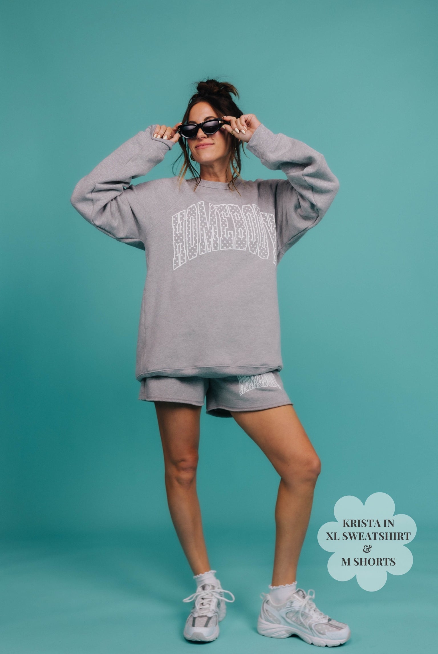 Woman wearing a gray sweatshirt and shorts with sunglasses on a teal background