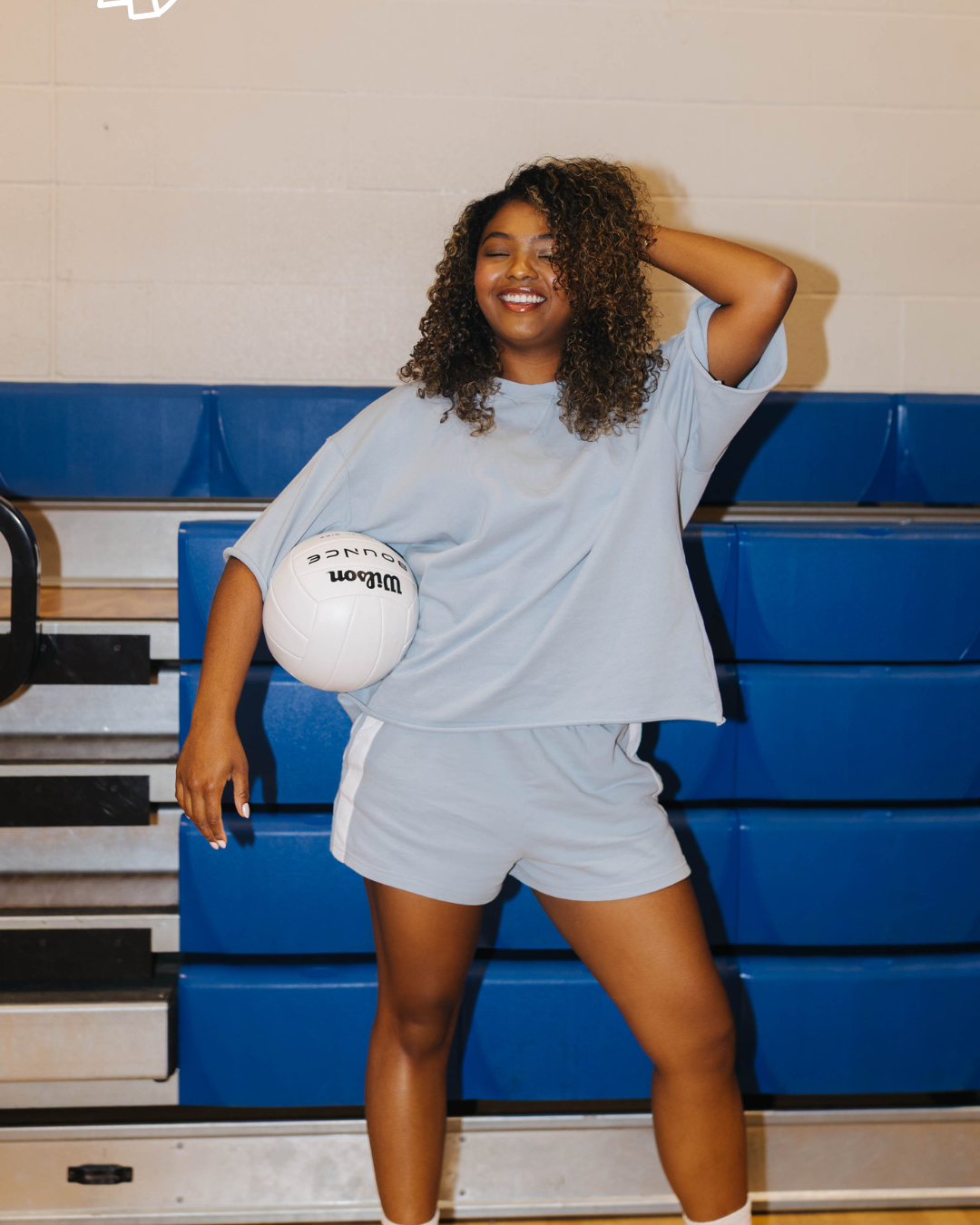 A woman posing in a gym wearing a light blue oversized t-shirt and matching shorts, holding a white ball.
