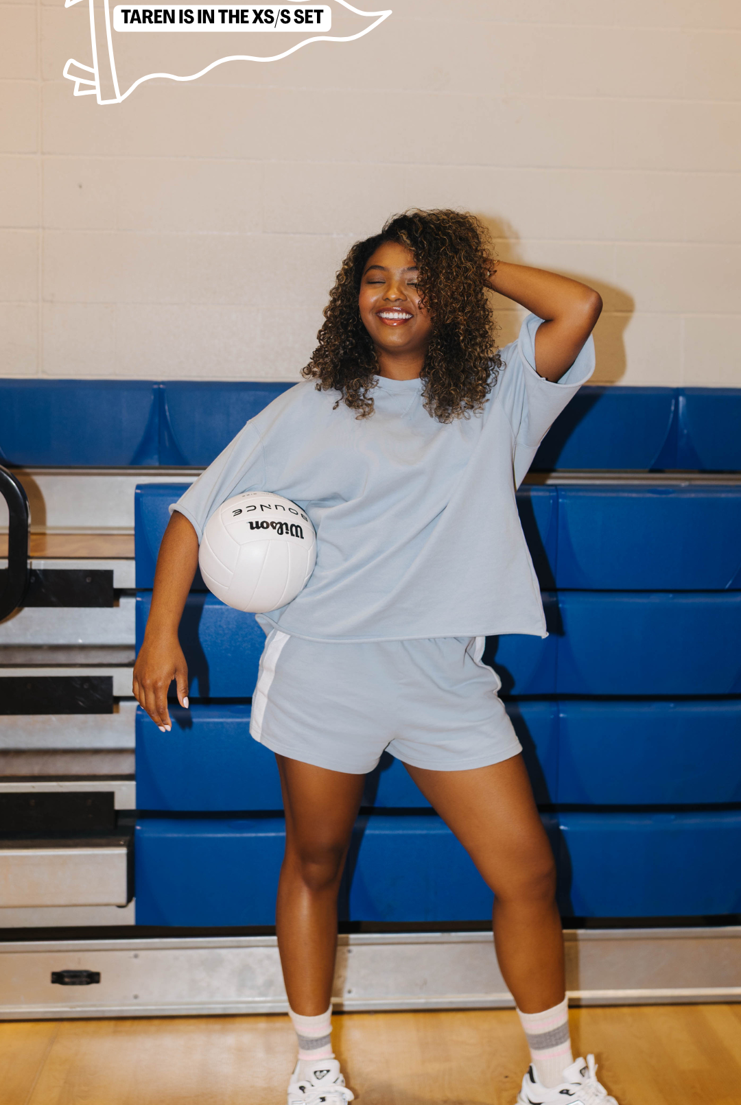 A woman posing in a gym wearing a light blue oversized t-shirt and matching shorts, holding a white ball.