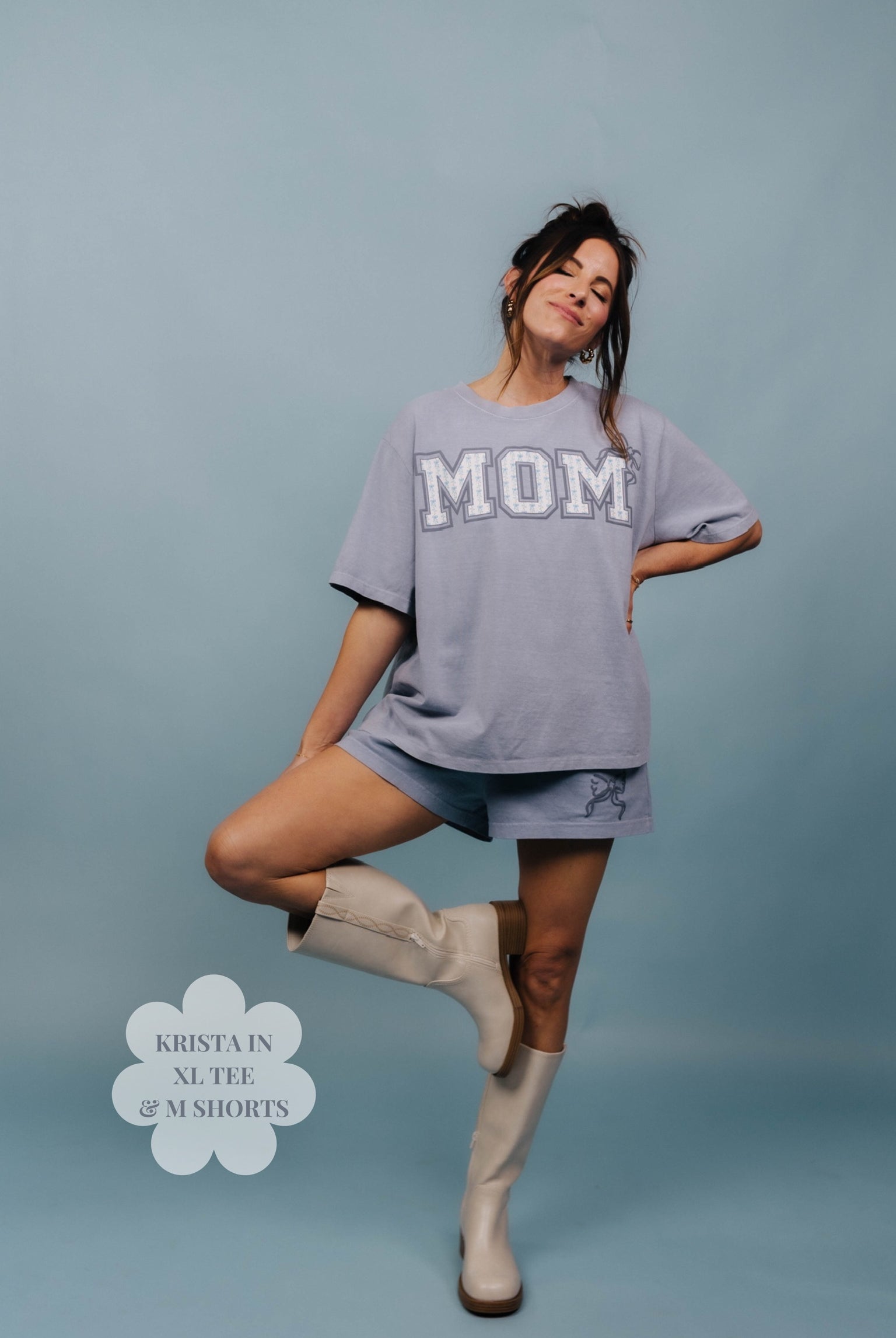Woman wearing a oversized 'MOM' shirt with shorts and boots on a blue background