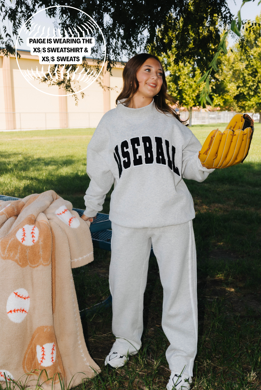 A woman standing outdoors on grass wearing a light gray sweatshirt and sweatpants set with 'BASEBALL' printed in black across the chest. She is holding a yellow baseball glove and a tan blanket with baseball patterns.