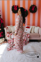 Woman in festive pajama set standing in a decorated room with Christmas wreaths and tree.