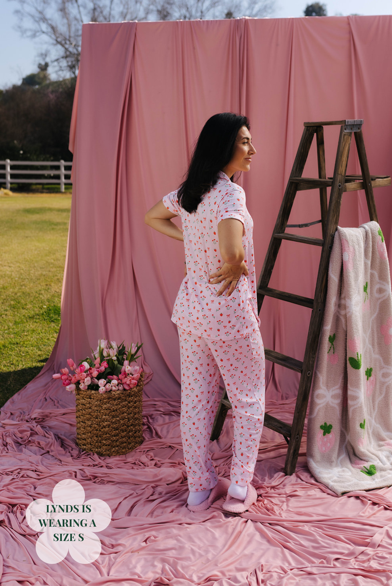 Woman in a floral pajama set standing in front of a pink backdrop with a ladder and flowers.