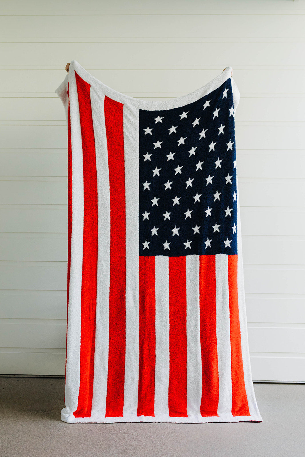 An American flag-themed blanket hanging against a white background, displaying red and white stripes with a blue field and white stars.