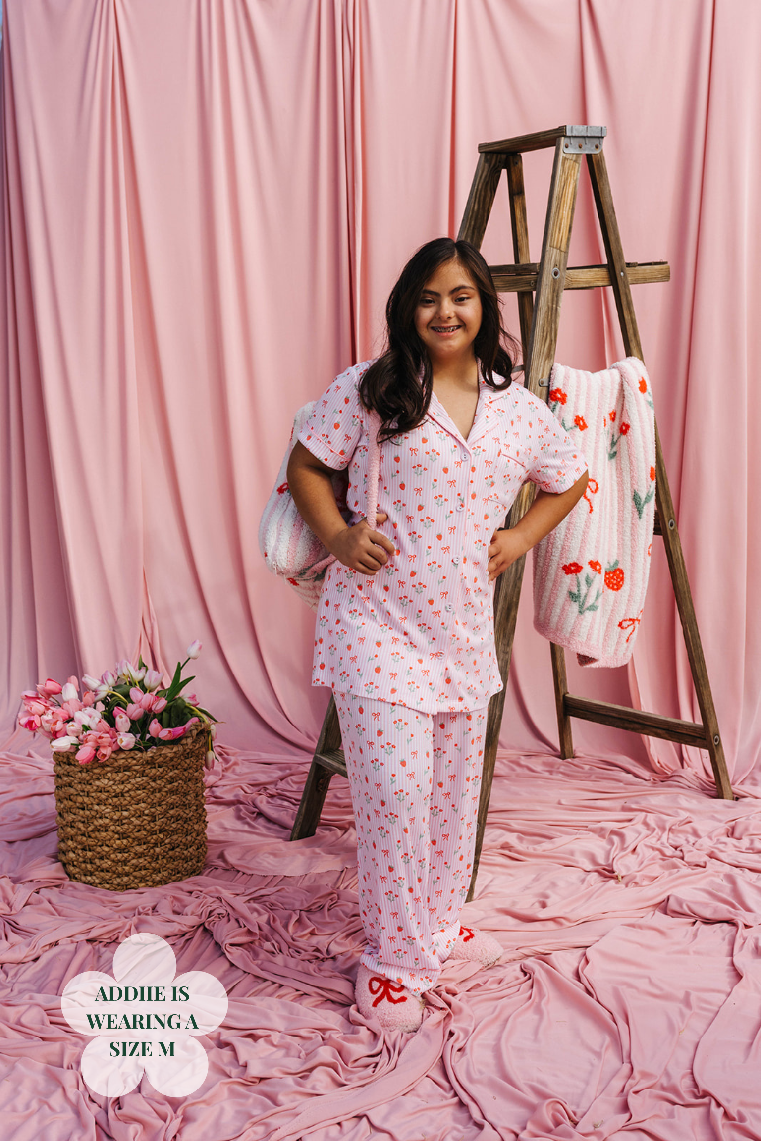 Woman wearing a pink floral pajama set standing next to a wooden ladder with more pajamas on it, all against a pink curtain background.