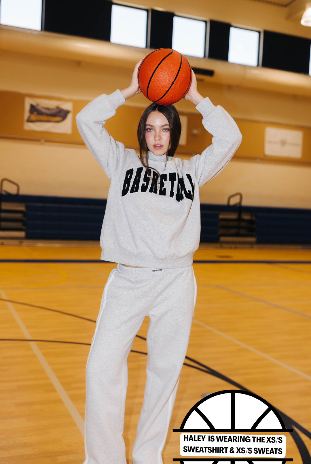 A woman stands on a basketball court wearing a light gray sweatshirt and sweatpants set with black 'BASKETBALL' lettering across the chest. She is holding an orange basketball above her head.