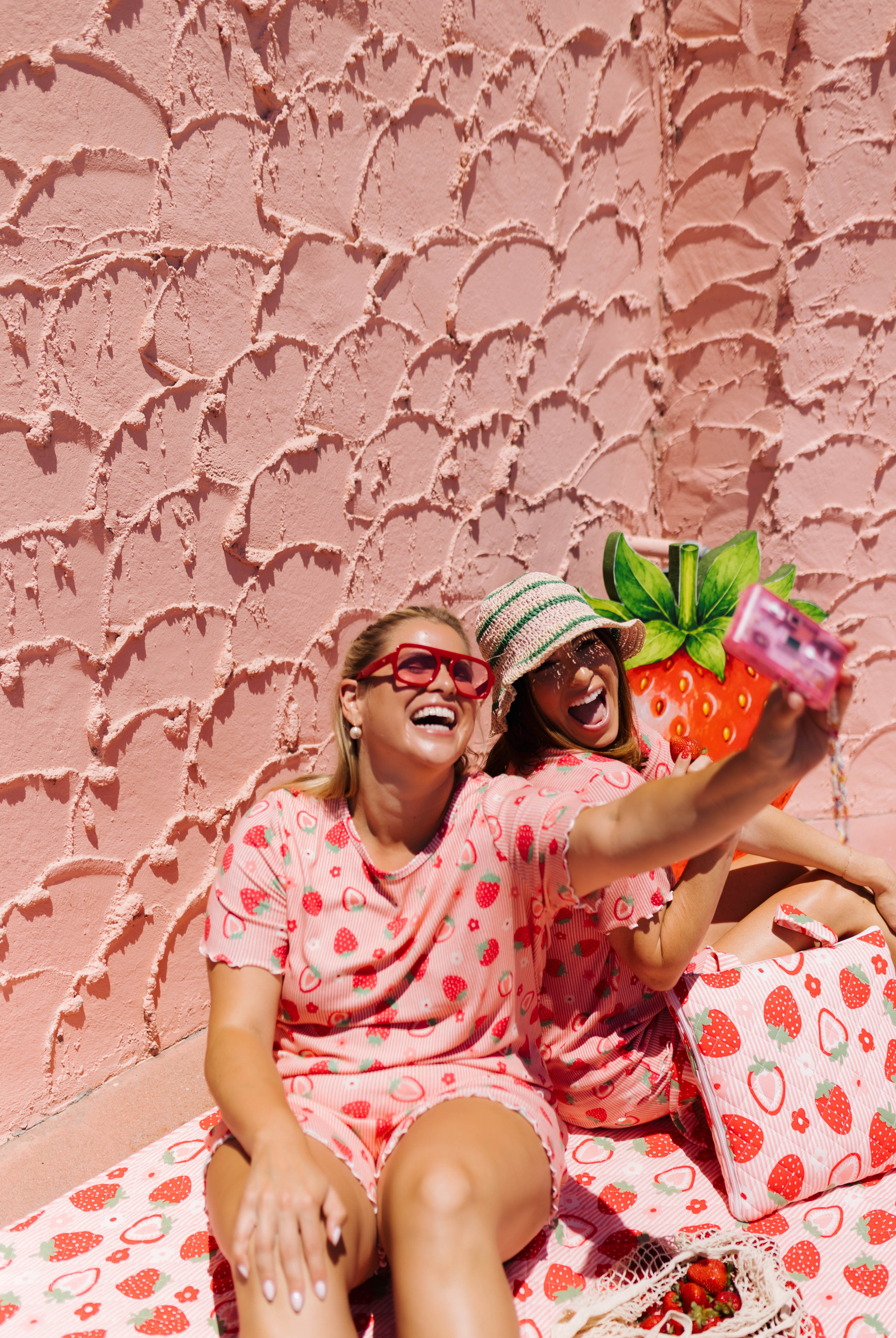 Two women wearing matching pink strawberry-print pajamas take a selfie in front of a textured pink wall.