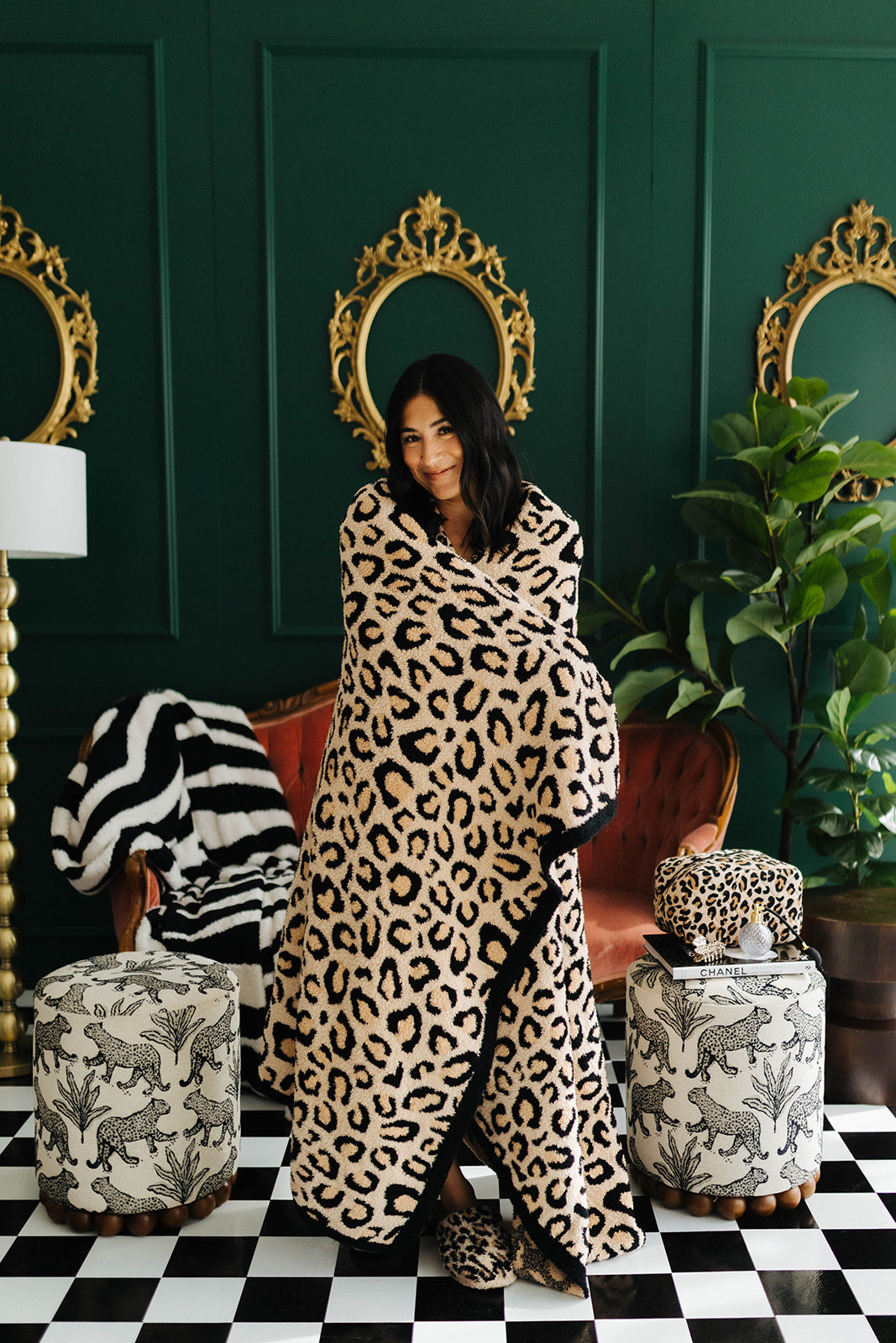 Woman wrapped in a leopard print blanket in a stylish room with green walls, gold mirrors, and patterned furniture.