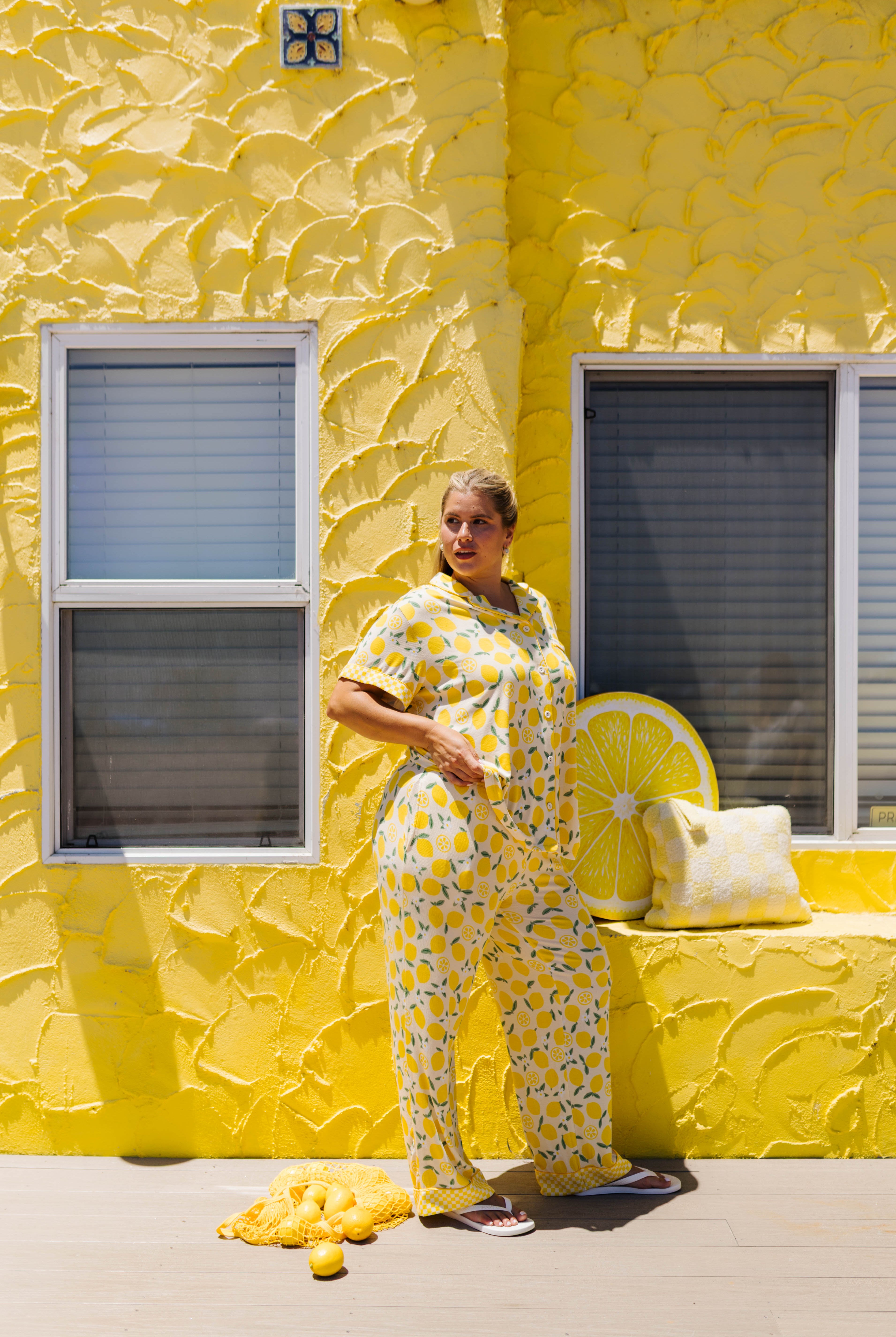 A woman stands outdoors against a bright yellow textured wall wearing a matching yellow and lemon-print pajama set.
