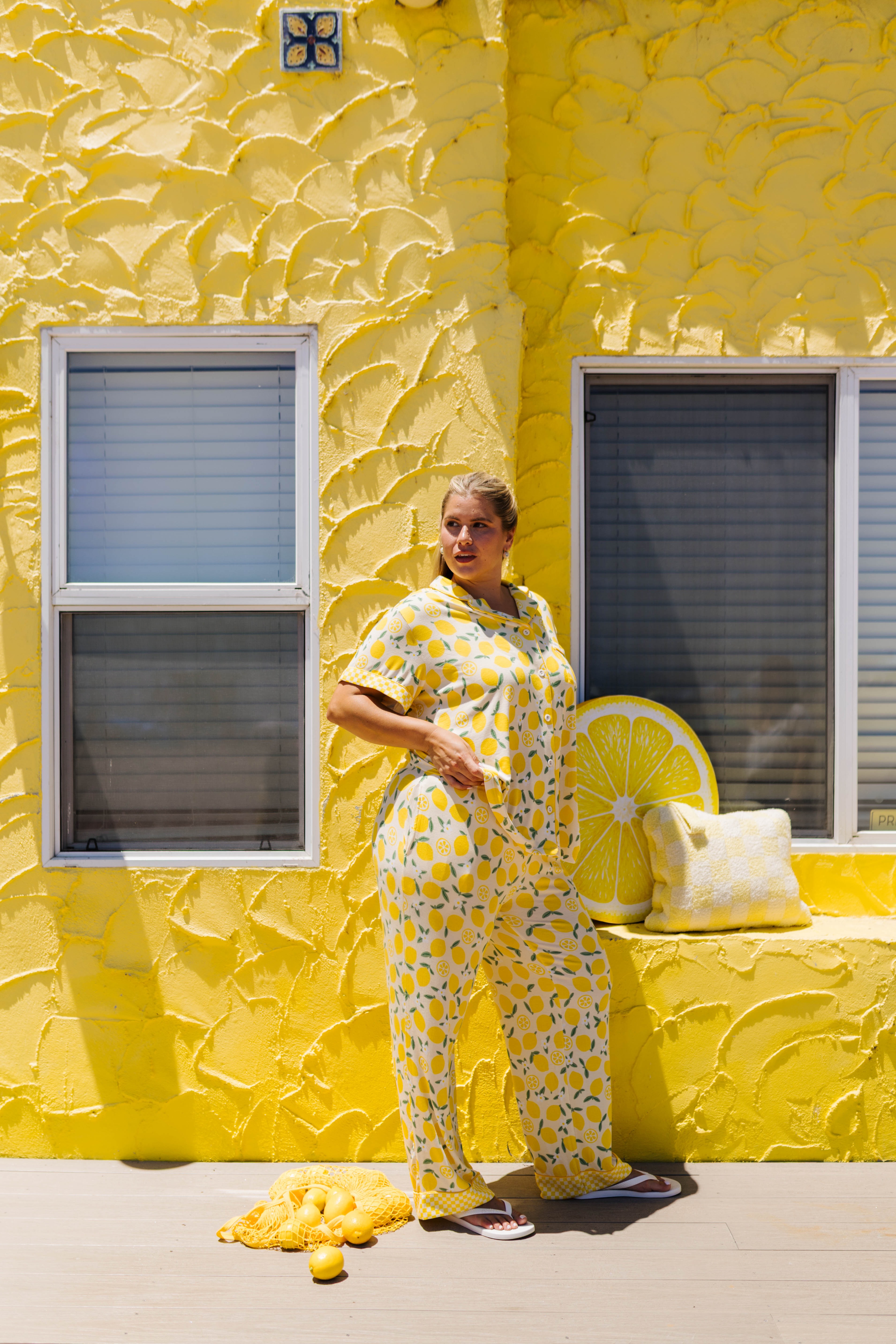 A woman stands outdoors against a bright yellow textured wall wearing a matching yellow and lemon-print pajama set.