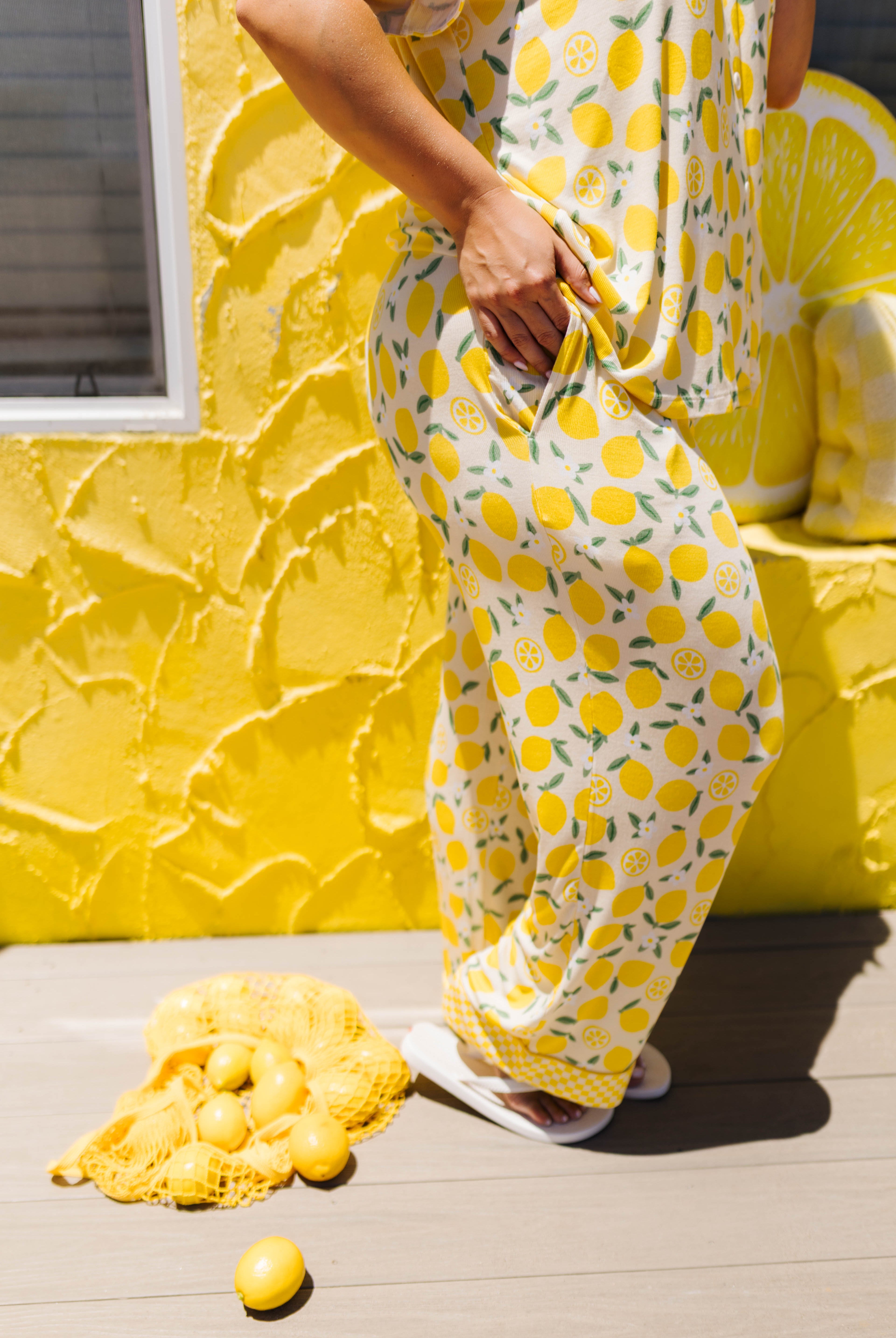 A person wearing a yellow lemon-print pj, standing on a light deck in front of a bright yellow textured wall with lemon-themed decor.