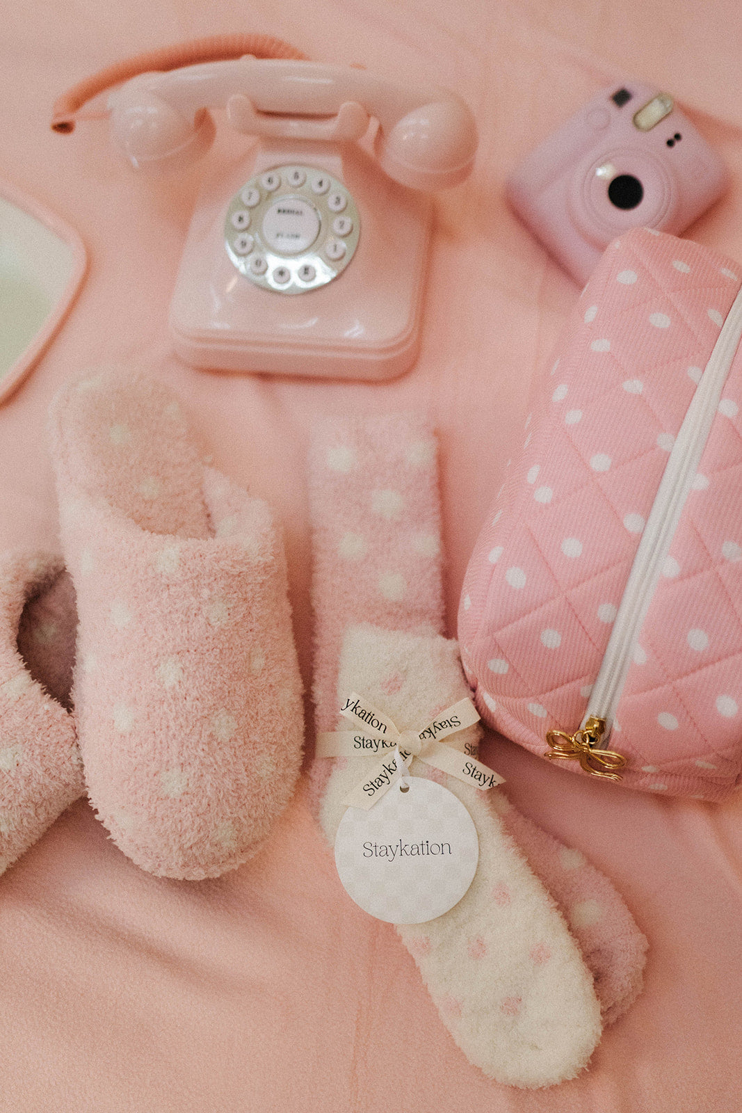 Collection of pink and white items including a phone, camera, pouch, and slippers on a pink background.