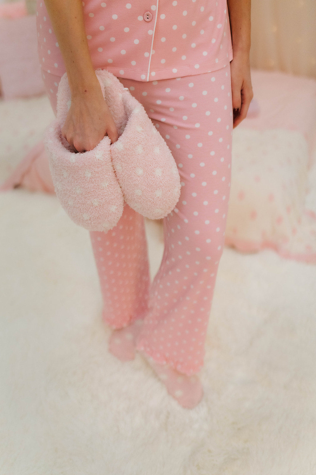 Person wearing pink polka dot pajamas holding pink slippers on a white surface.