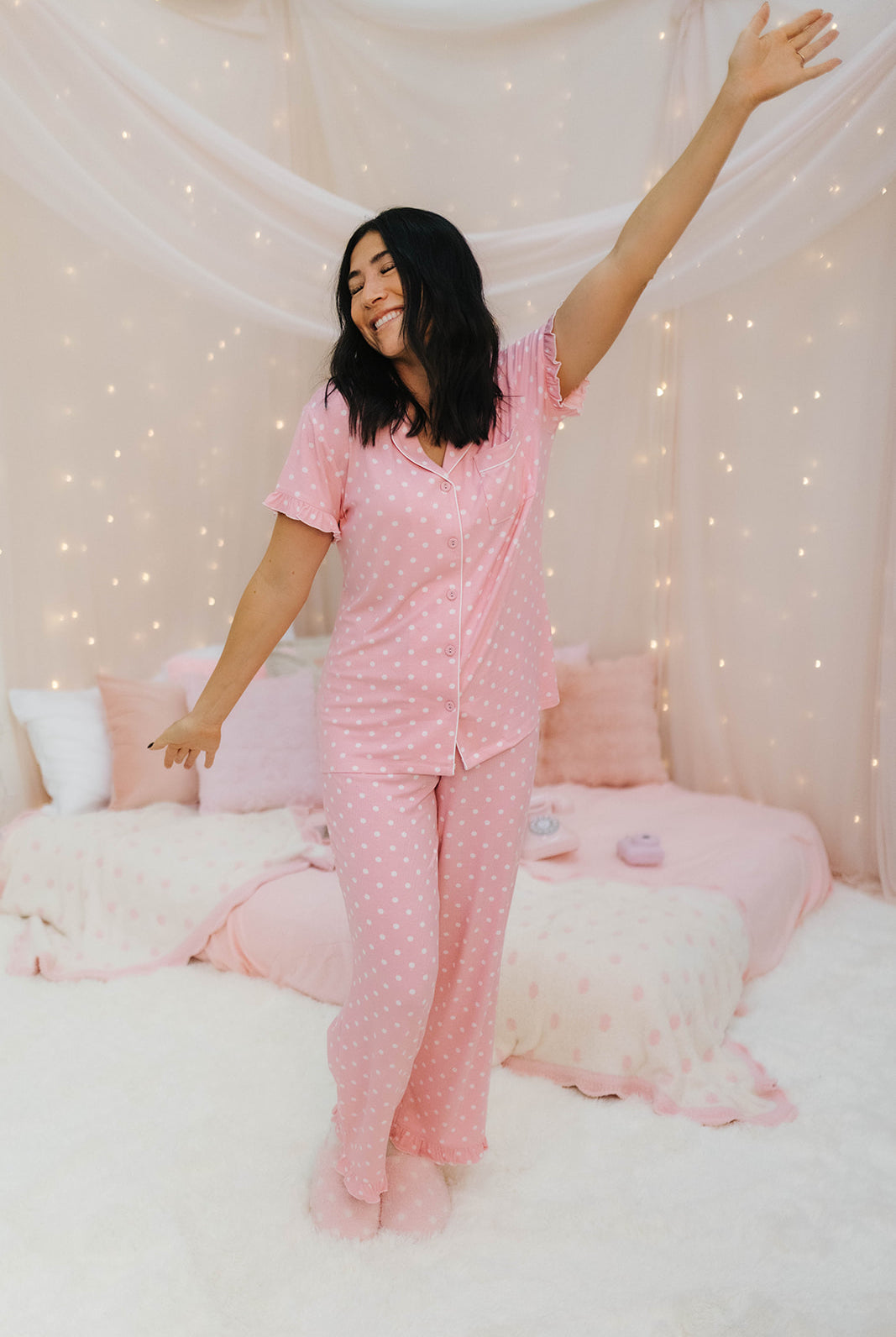 Woman in pink polka dot pajamas standing in a bedroom with white walls and decorative lights.