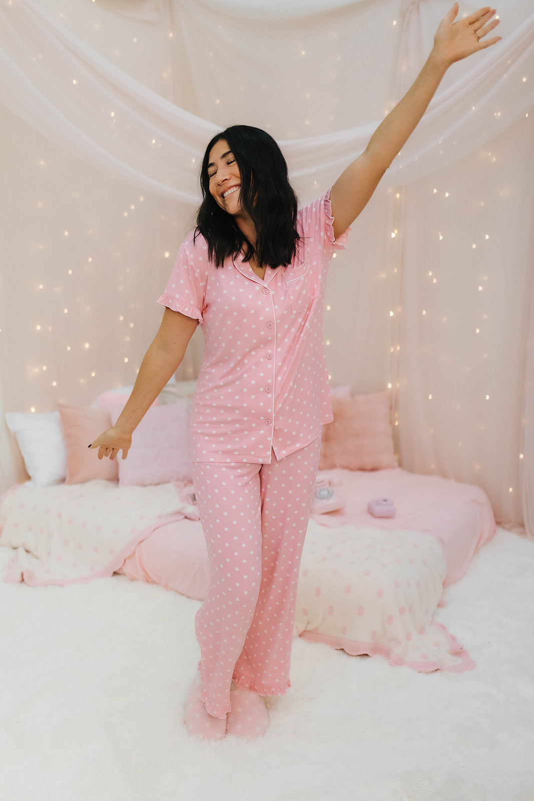 Woman in pink polka dot pajamas standing in a bedroom with white walls and decorative lights.