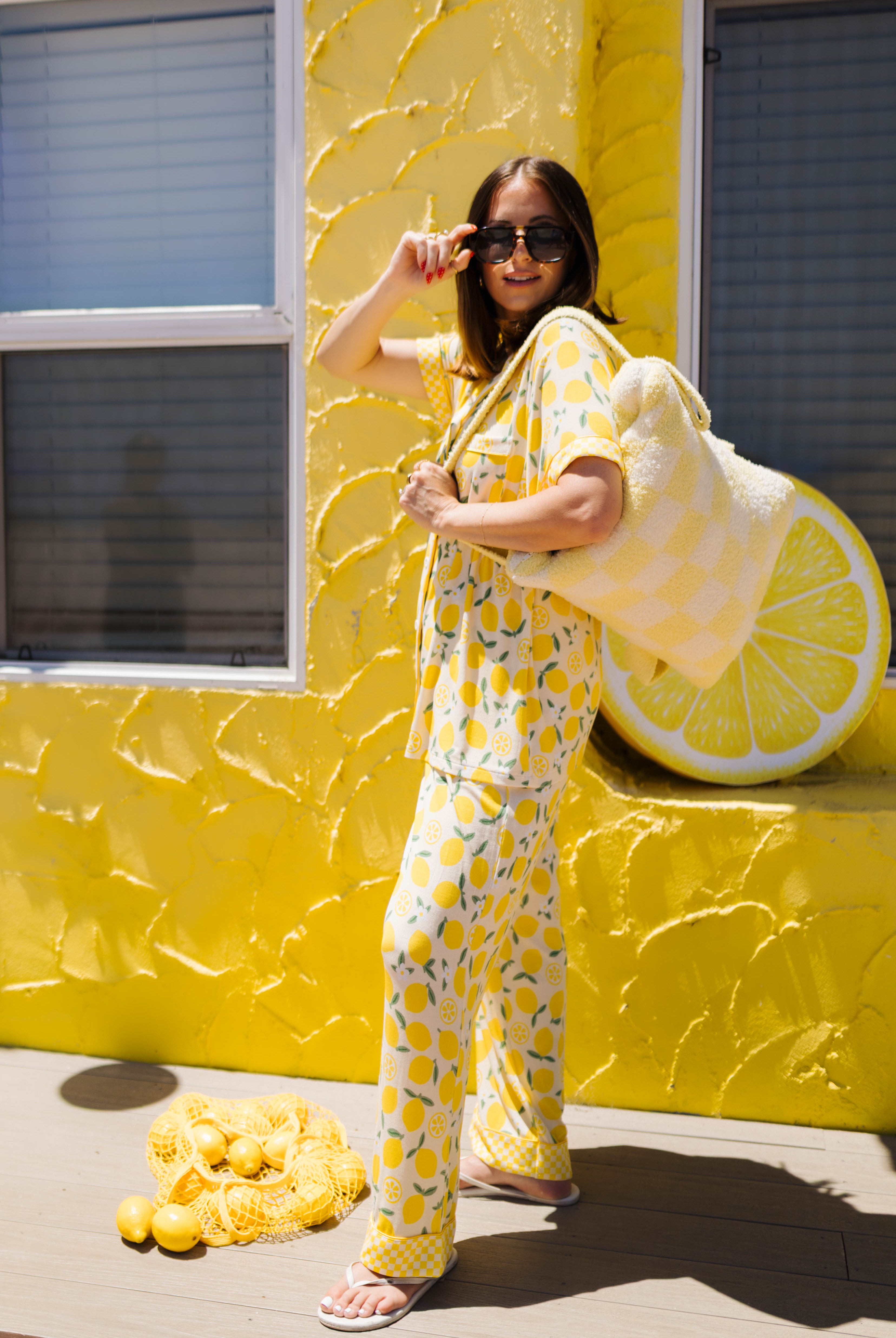 A woman wearing a yellow lemon-patterned pajama set stands in front of a bright yellow textured wall, holding a large plush quillow. A pile of lemons sits on the ground nearby.