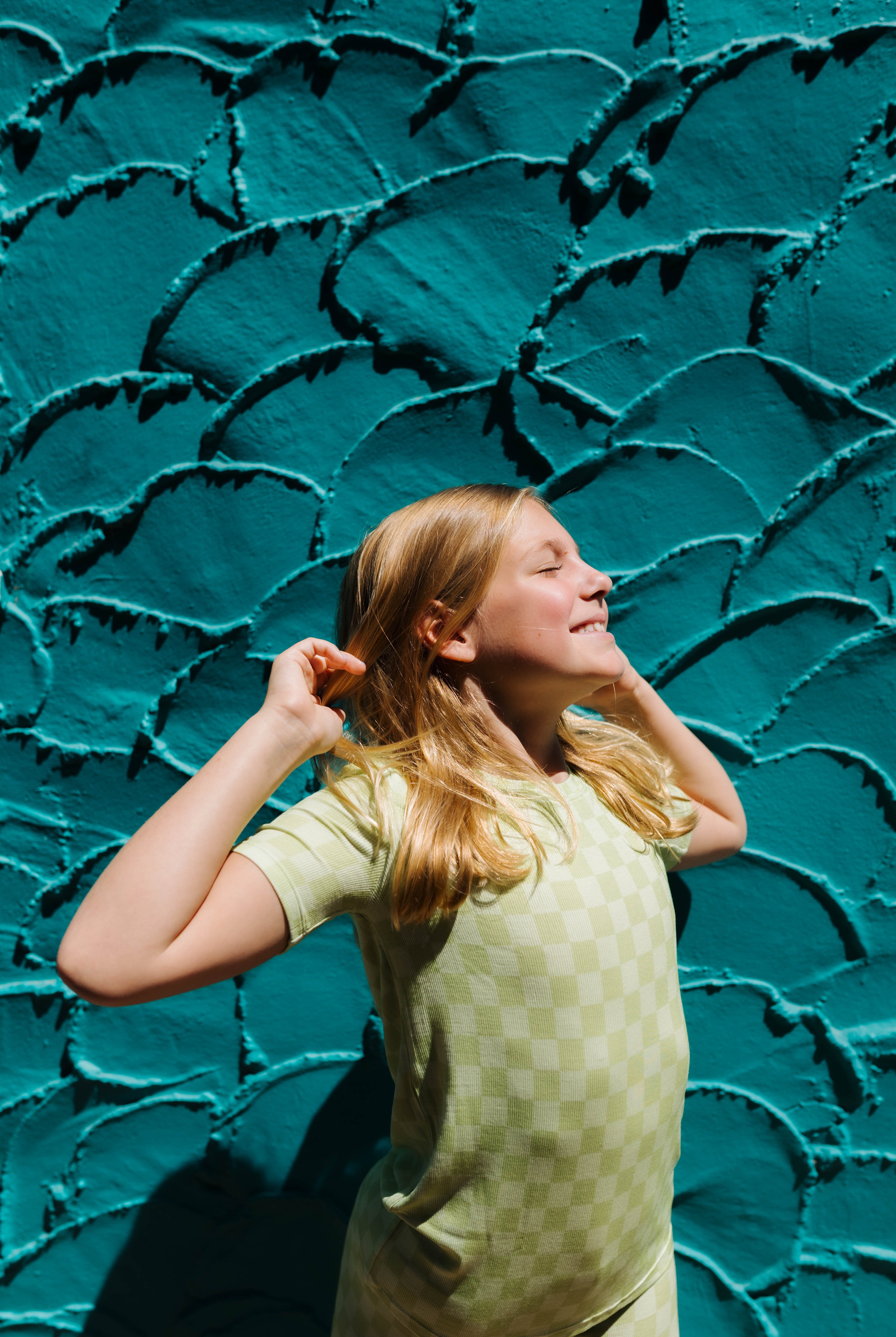 A young girl with blonde hair wearing a light green short sleeve top and matching pants, posing in front of a textured teal wall.