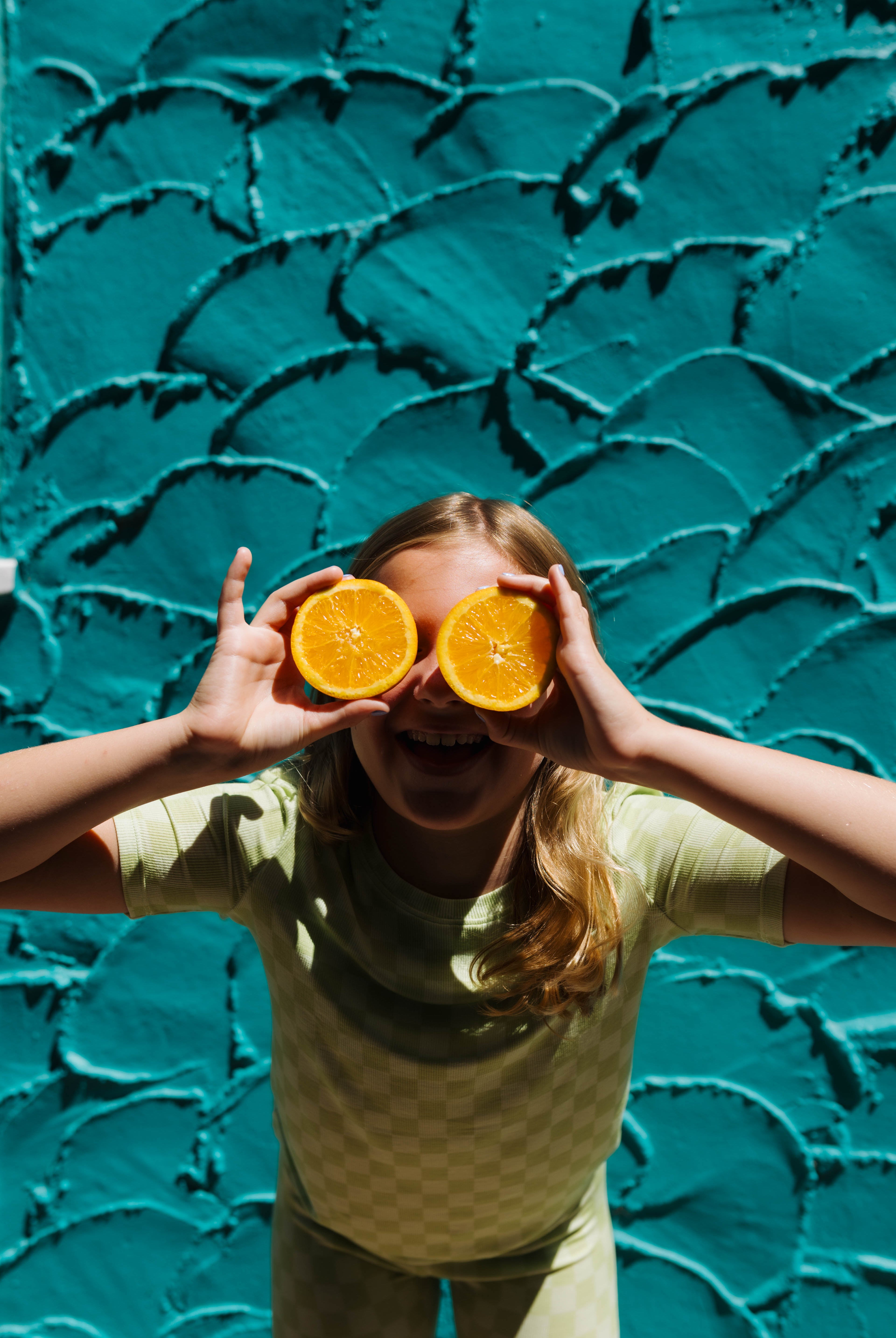 A young girl with blonde hair wearing a light green check pjs holds two orange halves up to her eyes, posing playfully in front of a textured teal wall.