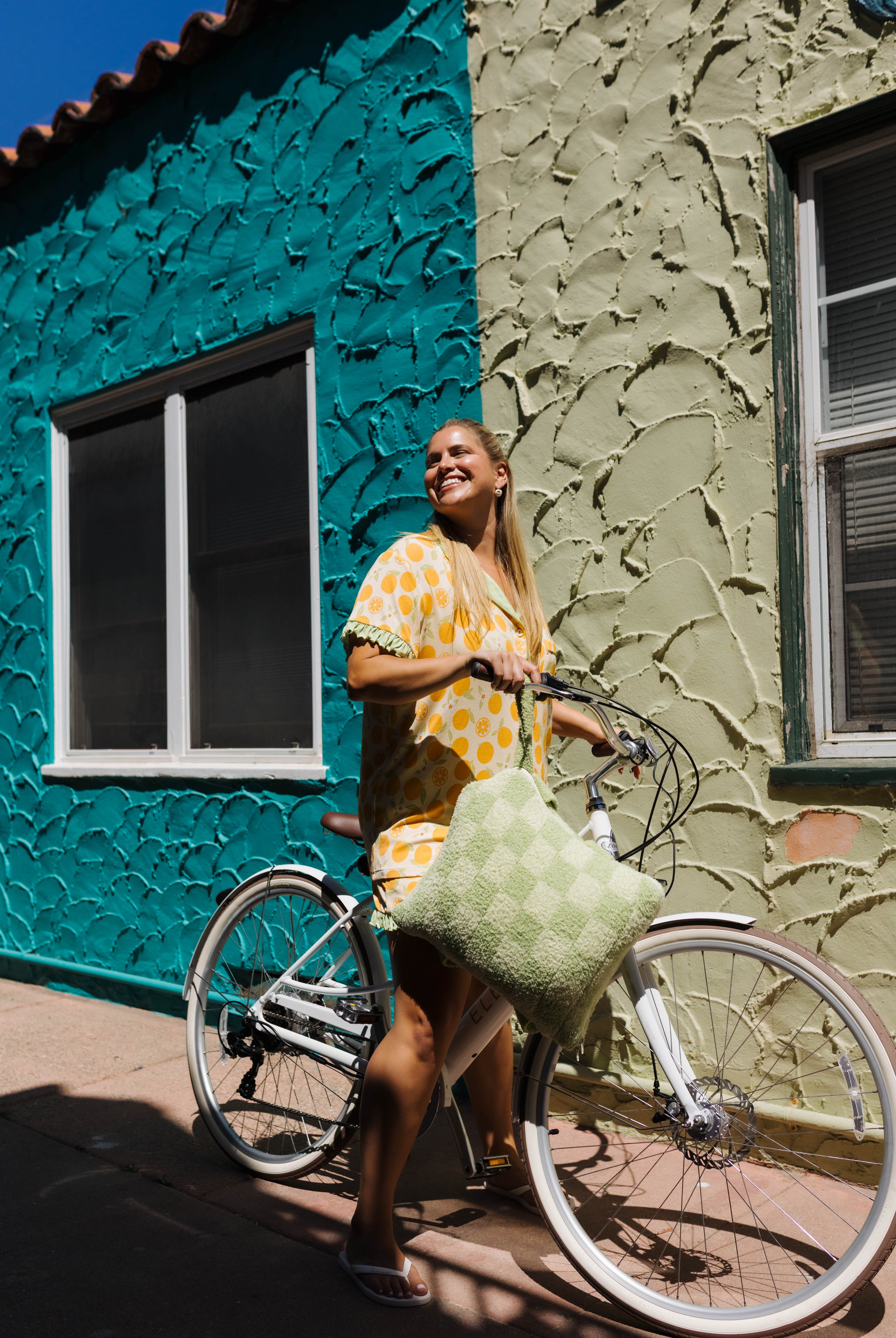 A woman smiling and riding a white bicycle outdoors next to a colorful textured building wall with a green check quillow