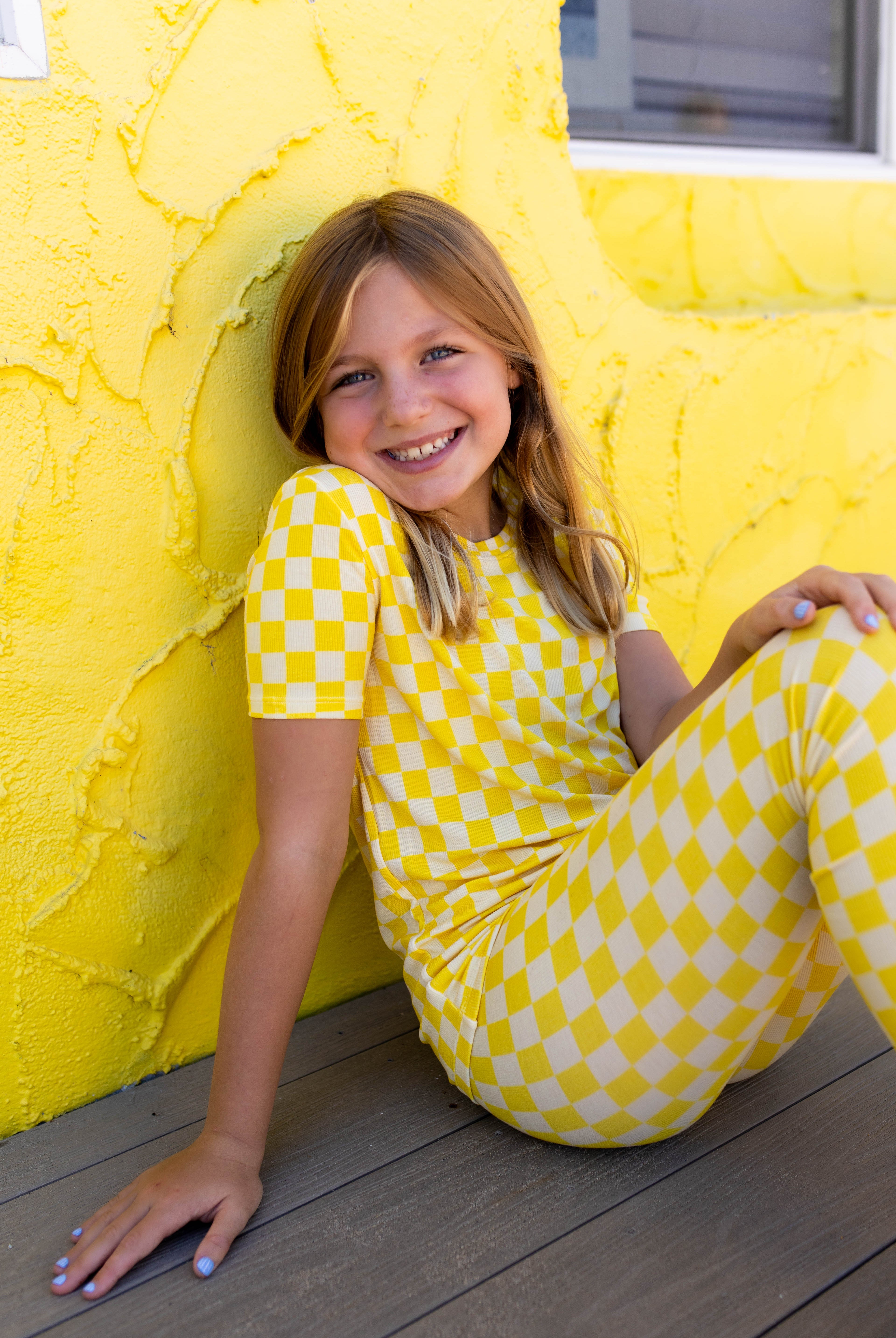 A young girl with blonde hair sits smiling on a wooden deck against a bright yellow stucco wall, wearing a matching yellow and checkered short-sleeve top pj set.