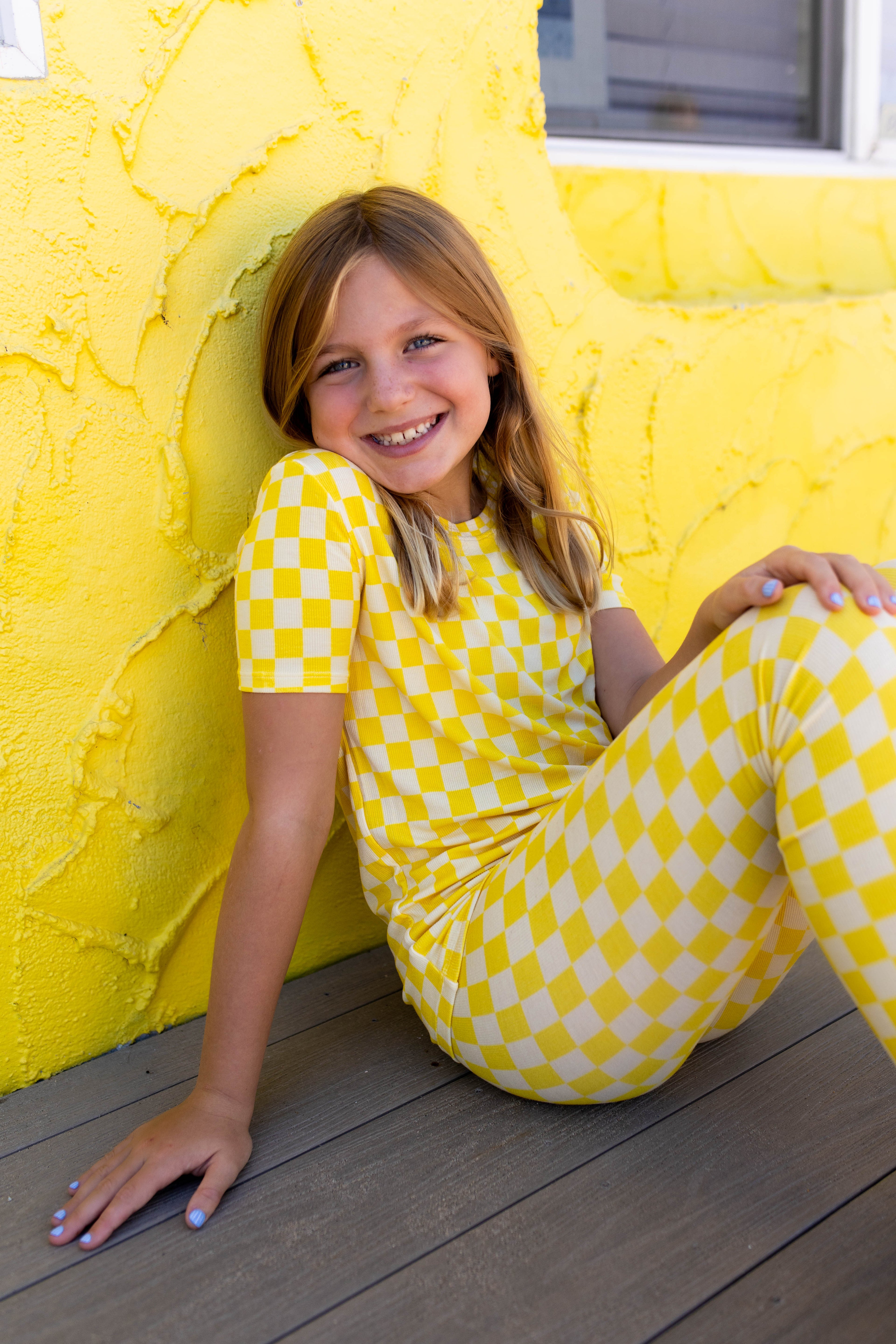 A young girl with blonde hair sits smiling on a wooden deck against a bright yellow stucco wall, wearing a matching yellow and checkered short-sleeve top pj set.