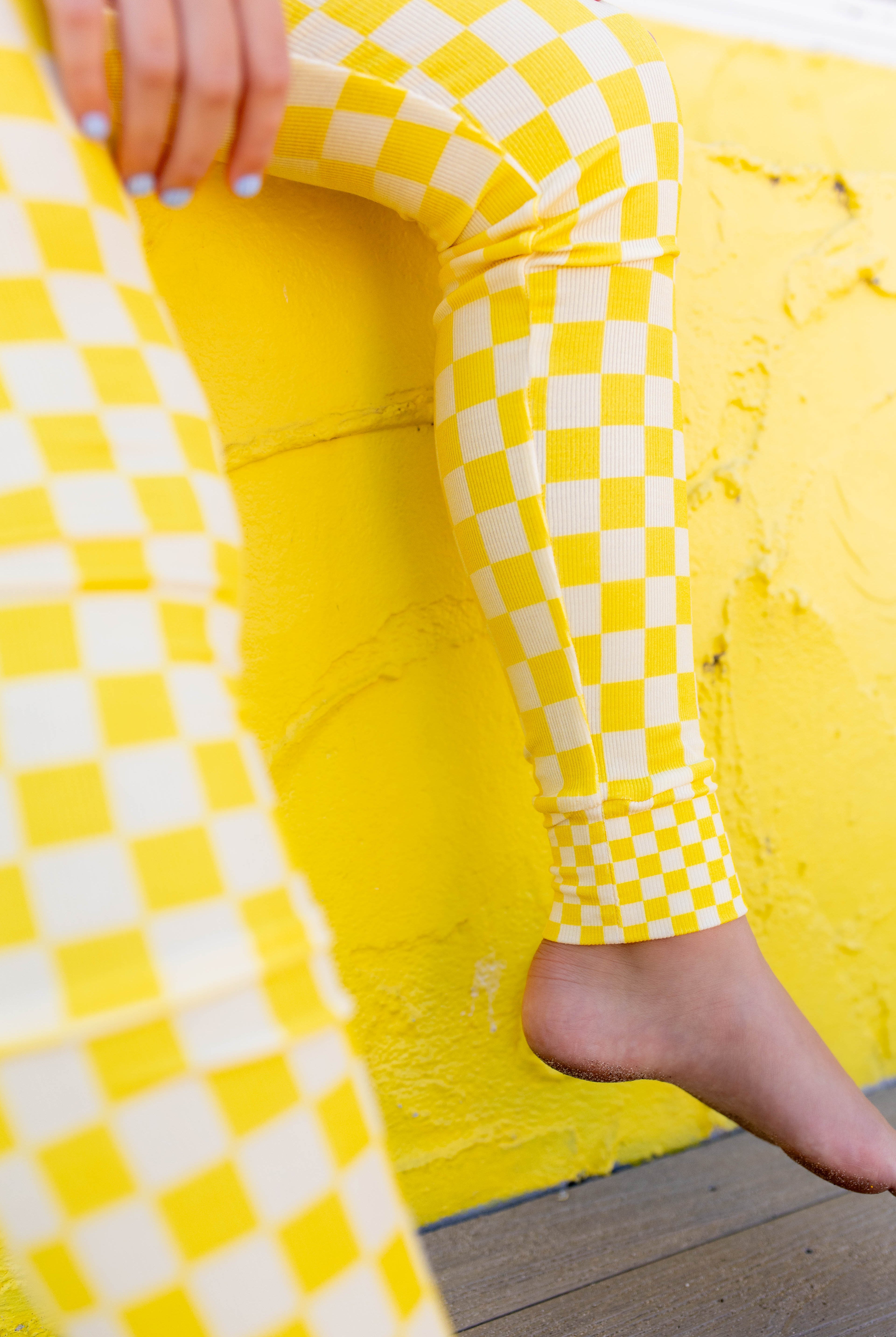 Close-up of a person wearing bright yellow and white checkerboard leggings, seated against a textured yellow wall with one bare foot visible.