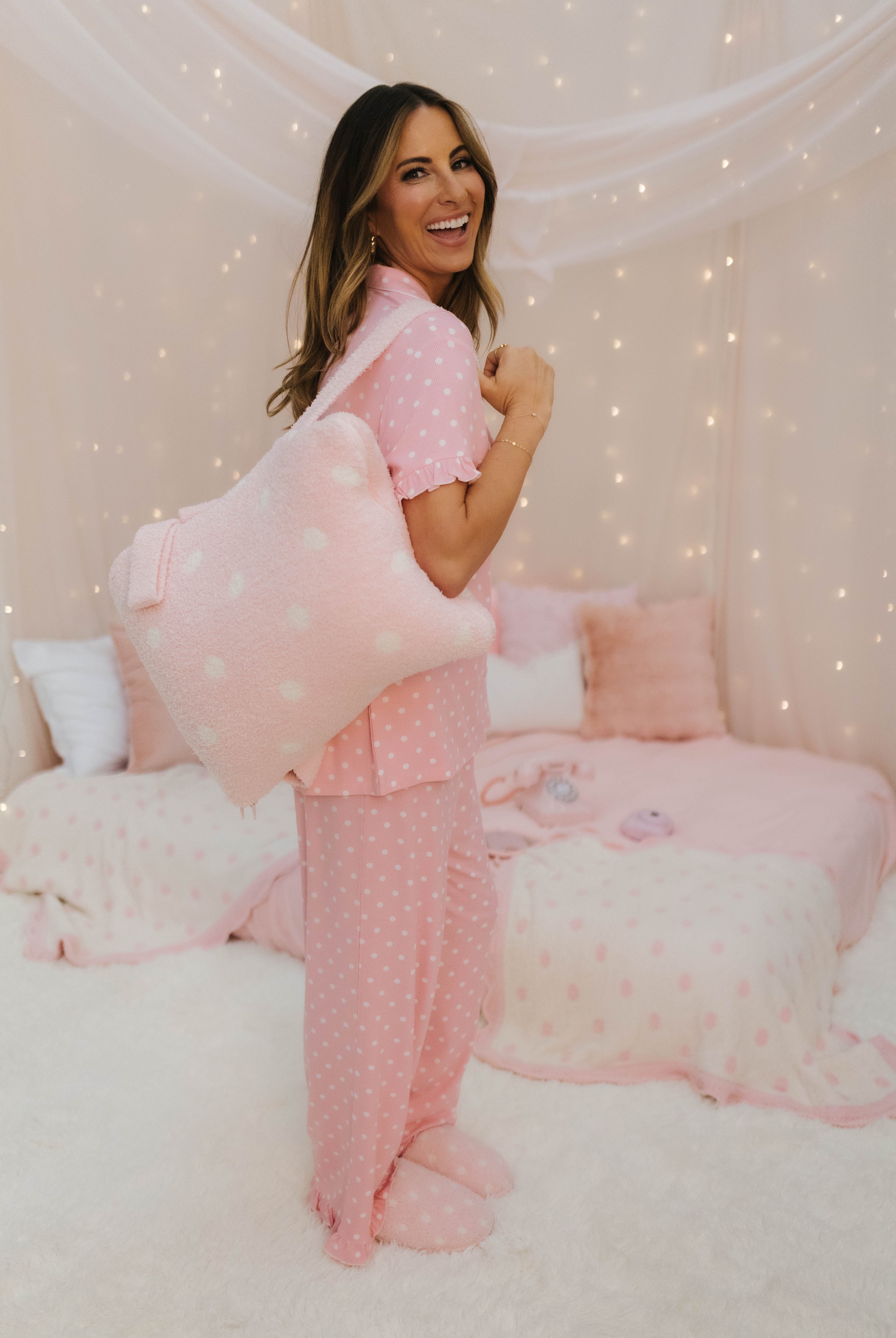 Woman in pink polka dot pajamas sitting on a bed with pink pillows and blankets.