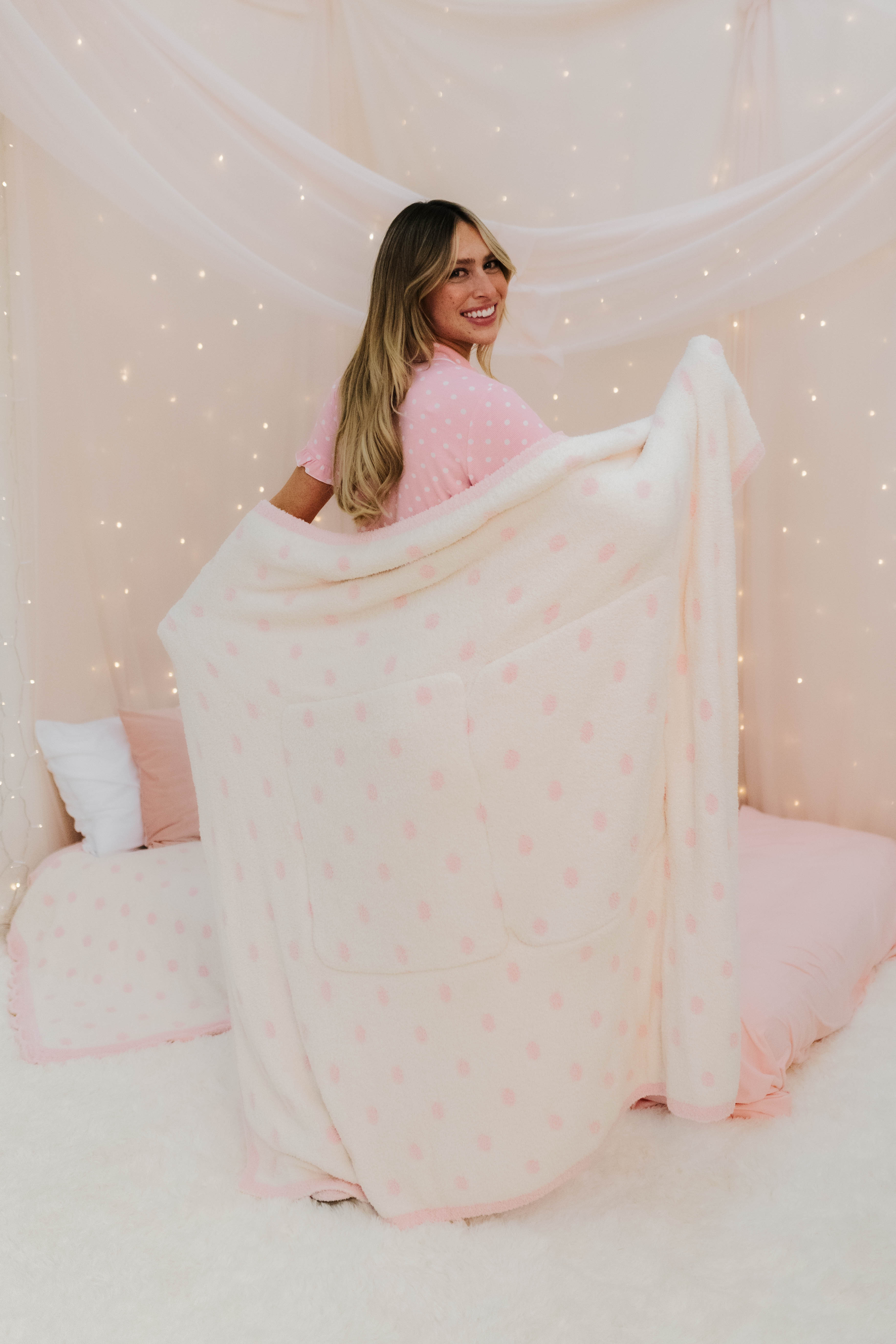 Woman in a pink outfit sitting under a polka dot blanket with a star pattern, surrounded by fairy lights.