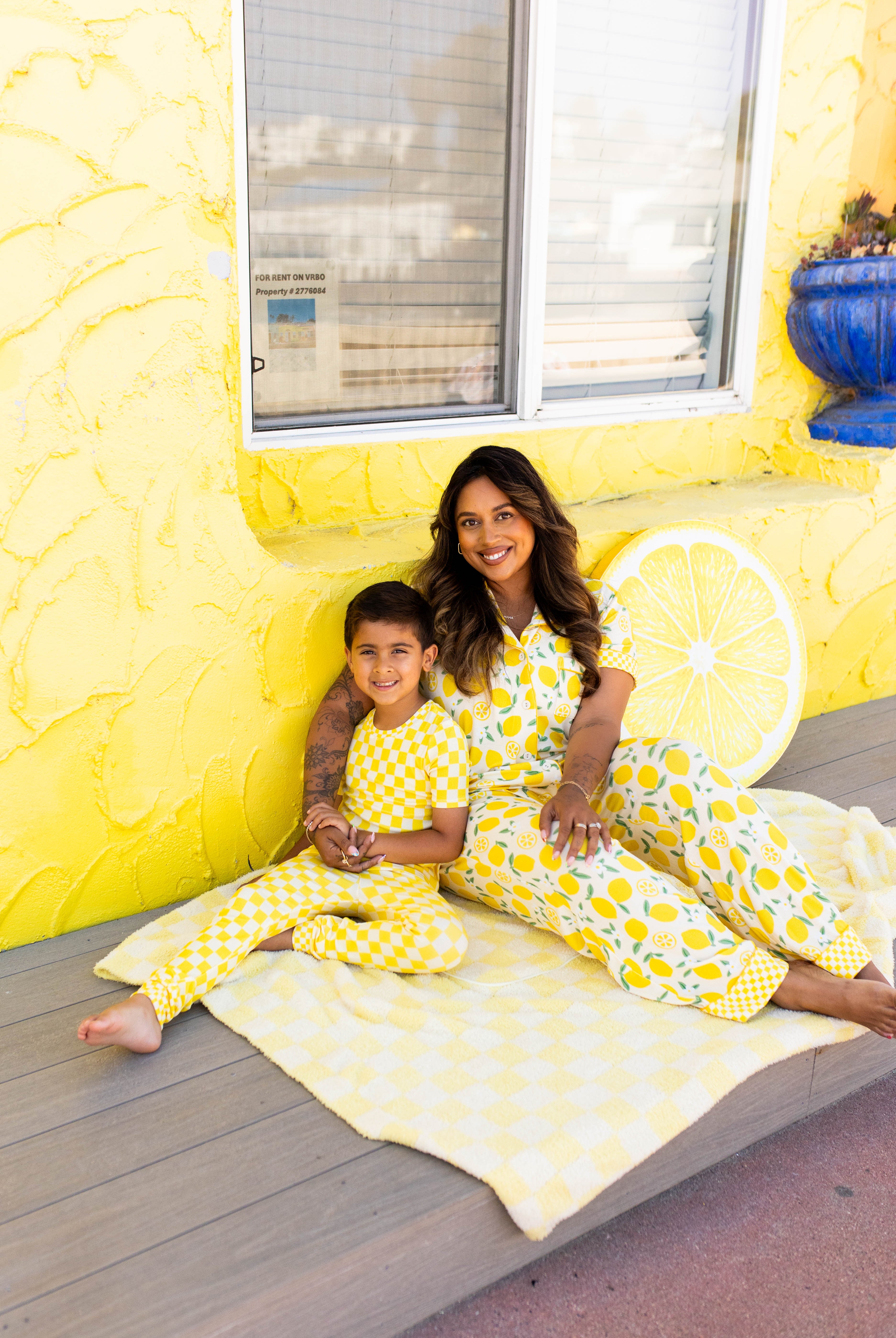 A woman and a young boy wearing coordinating pjs sit together on a light yellow blanket on a wooden deck in front of a bright yellow wall decorated with a lemon slice.