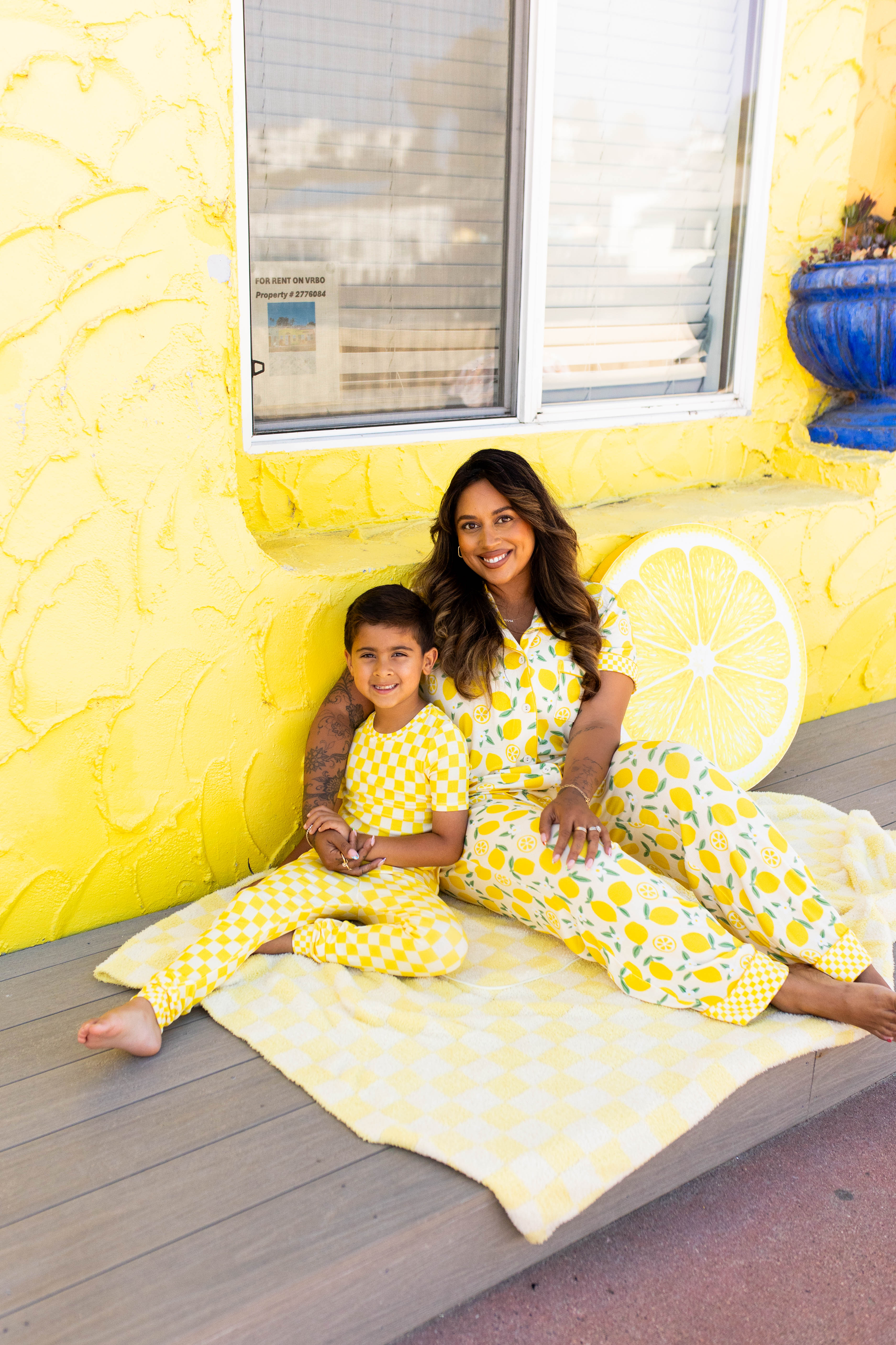 A woman and a young boy wearing coordinating pjs sit together on a light yellow blanket on a wooden deck in front of a bright yellow wall decorated with a lemon slice.