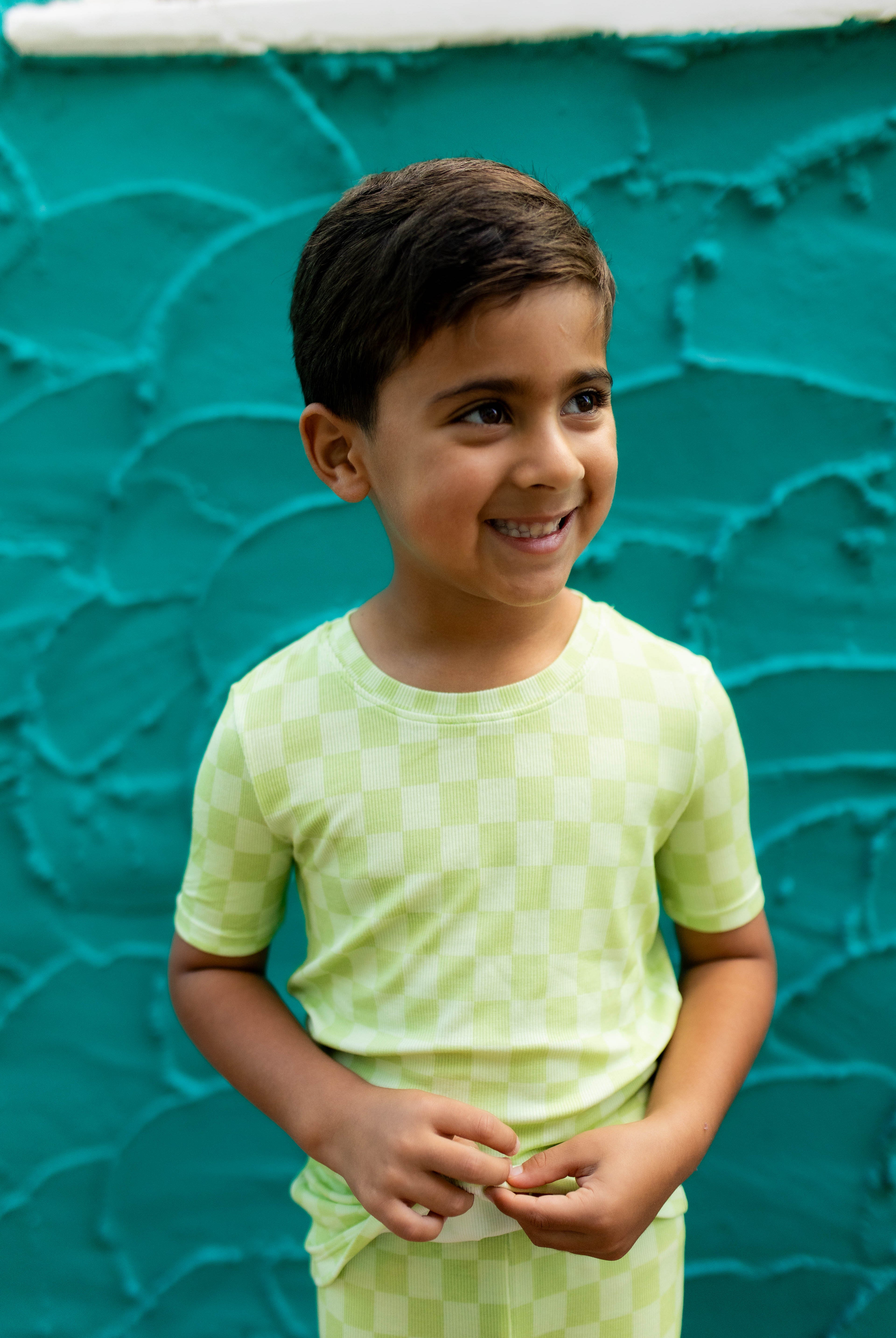 A young boy smiling and looking to the side, wearing a light green checkered short-sleeve t-shirt and matching pants, standing against a textured teal wall.