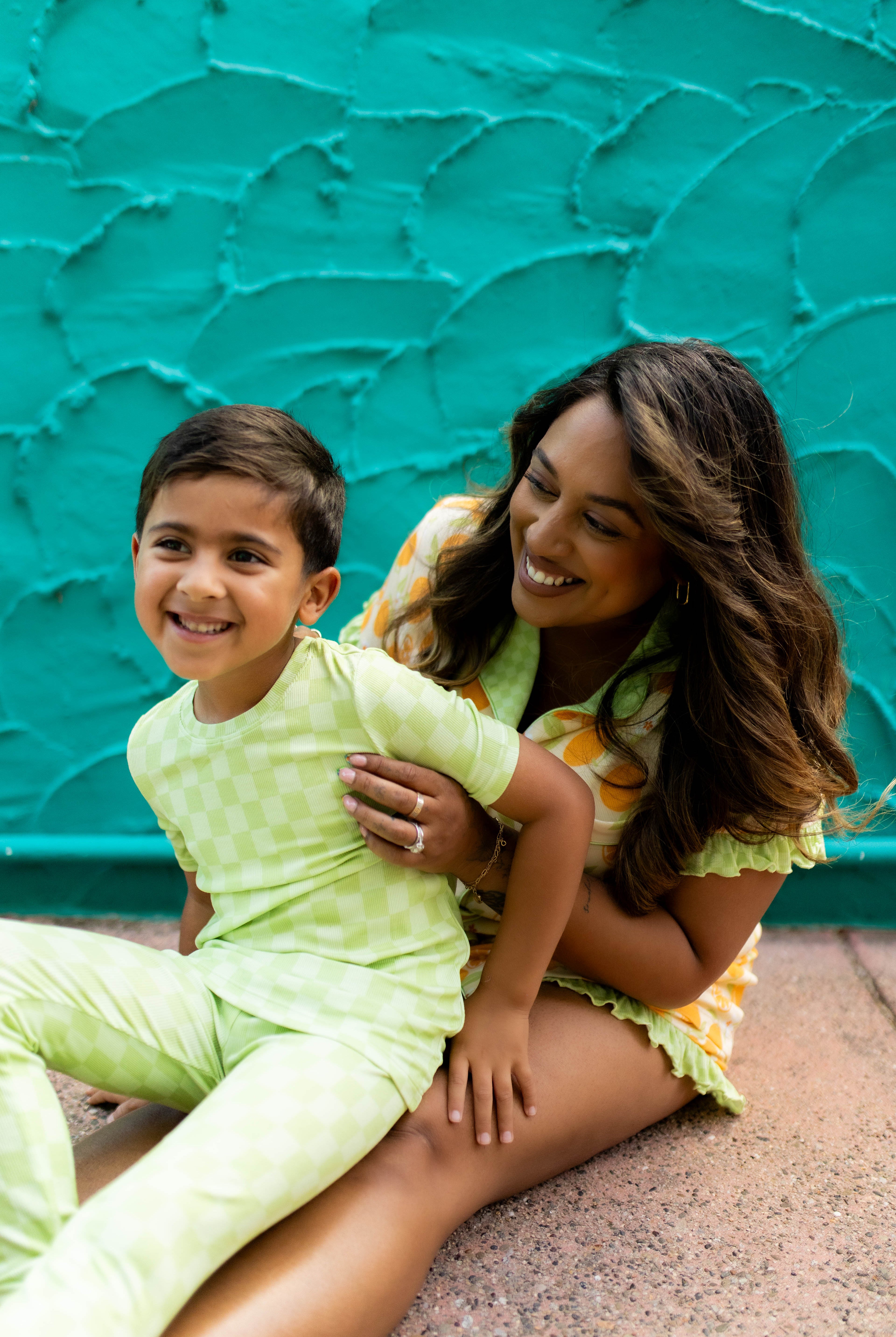 A woman and a young boy sitting together in front of a textured turquoise wall. The boy is wearing a light green checkered pajama set, and the woman is wearing a matching yellow and orange patterned pjs.
