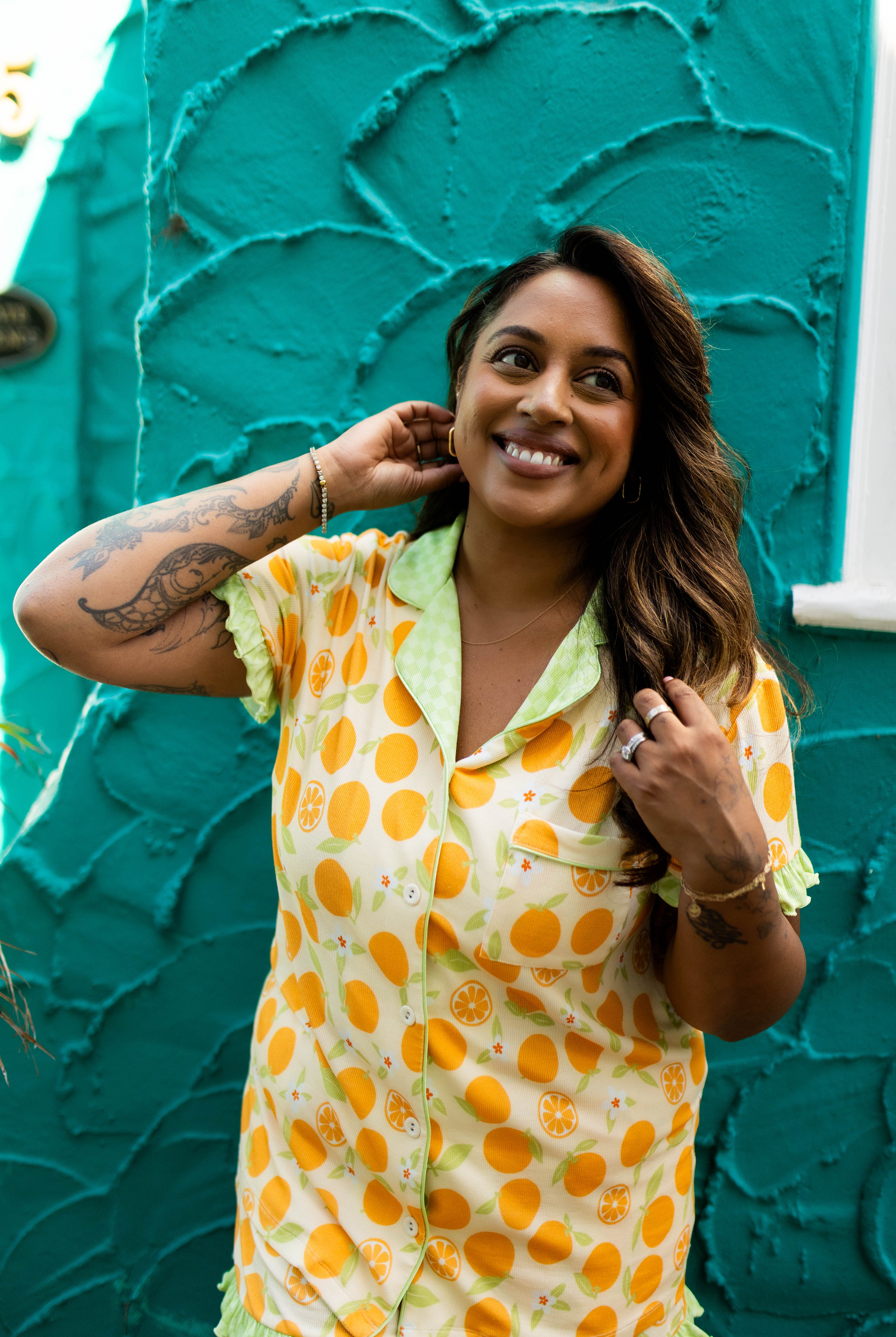 A woman smiling in a yellow and orange patterned short-sleeve pajama top with light green ruffled trim, standing in front of a textured teal wall.