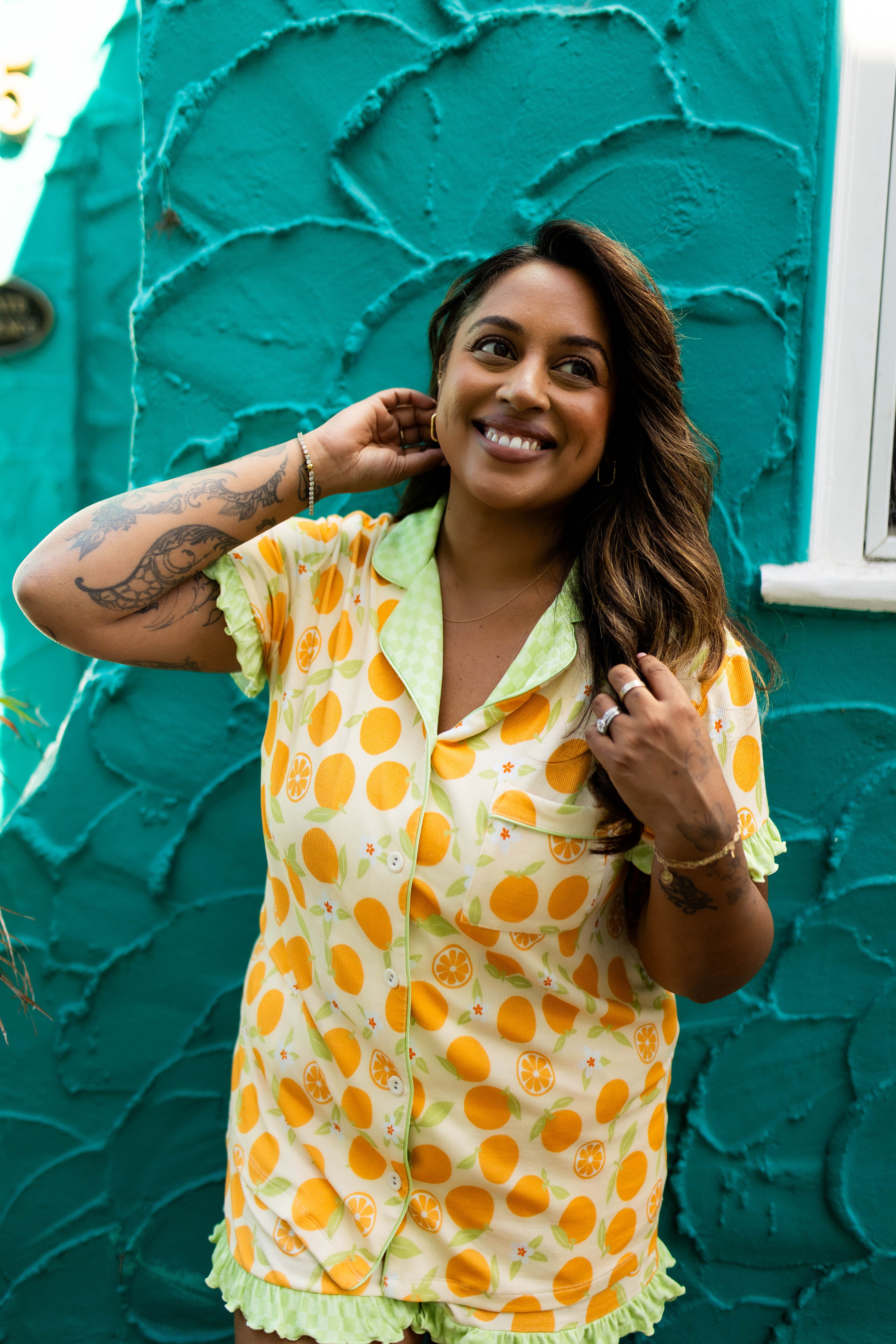 A woman smiling in a yellow and orange patterned short-sleeve pajama top with light green ruffled trim, standing in front of a textured teal wall.