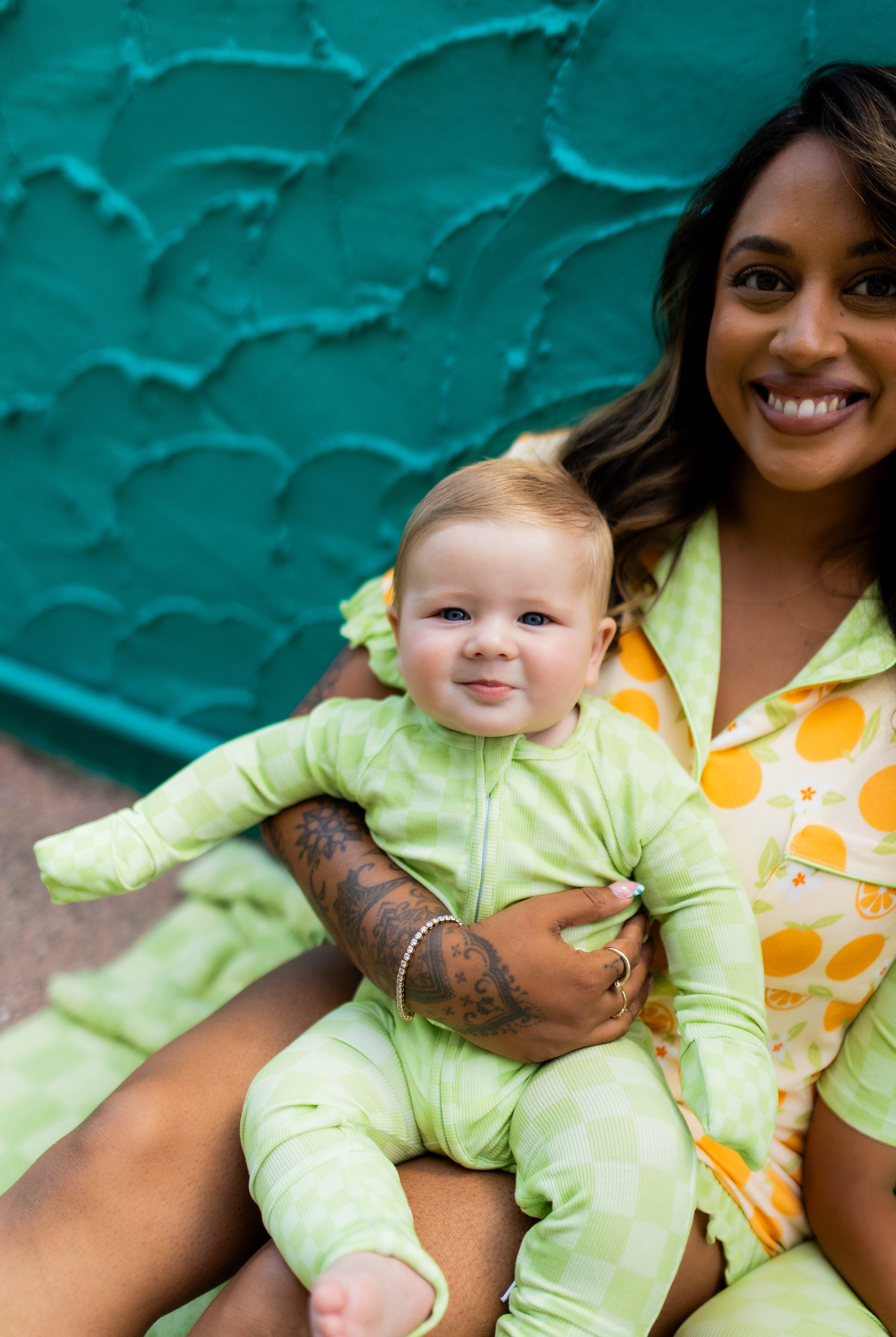 A smiling woman holding a baby wearing a light green footed pj, with the woman also wearing orange pjs, set against a textured teal wall.