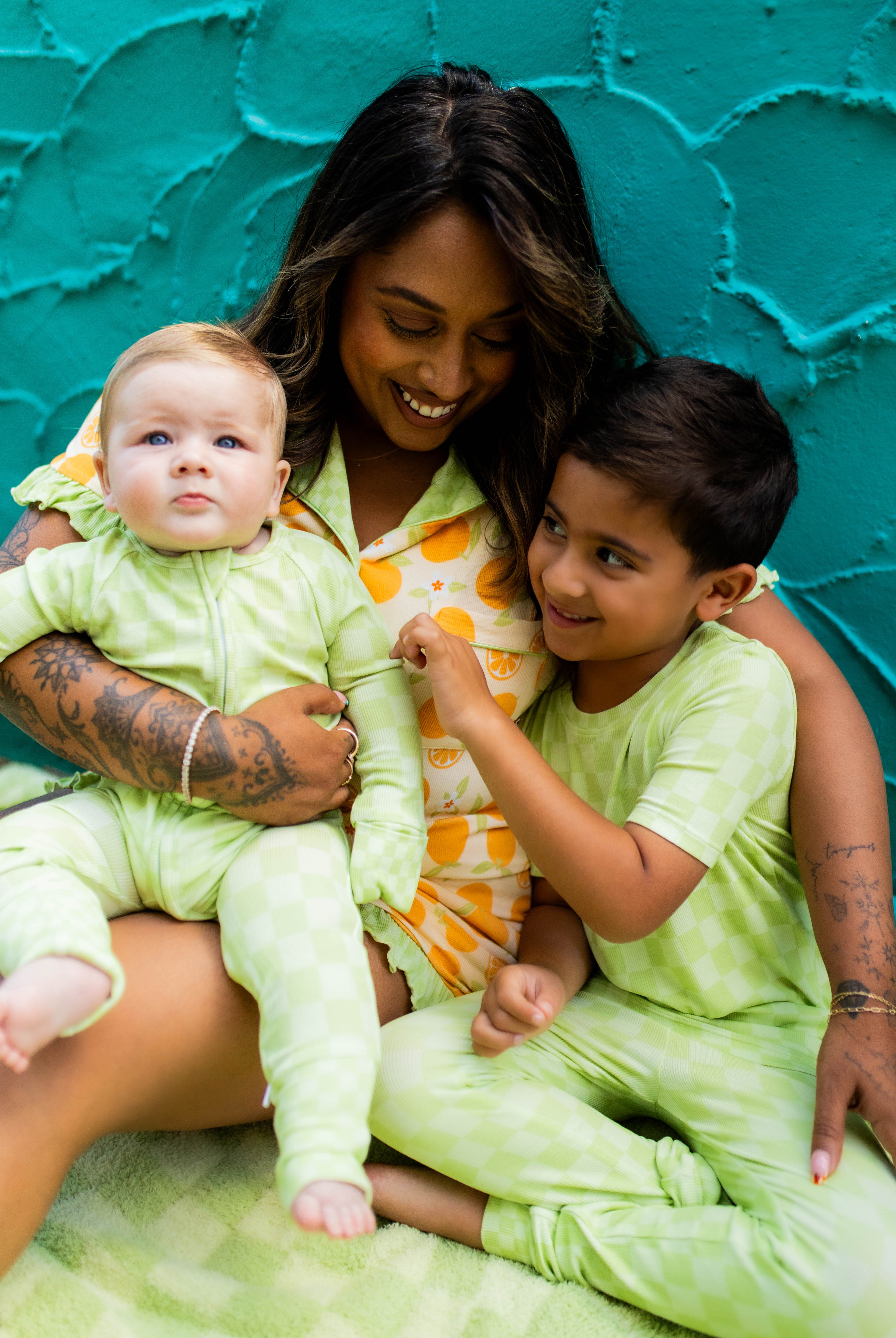 A woman and two children wearing matching light green pajamas sit together against a textured teal wall. The woman wears an orange patterned pajama top, while the children wear light green pajamas.
