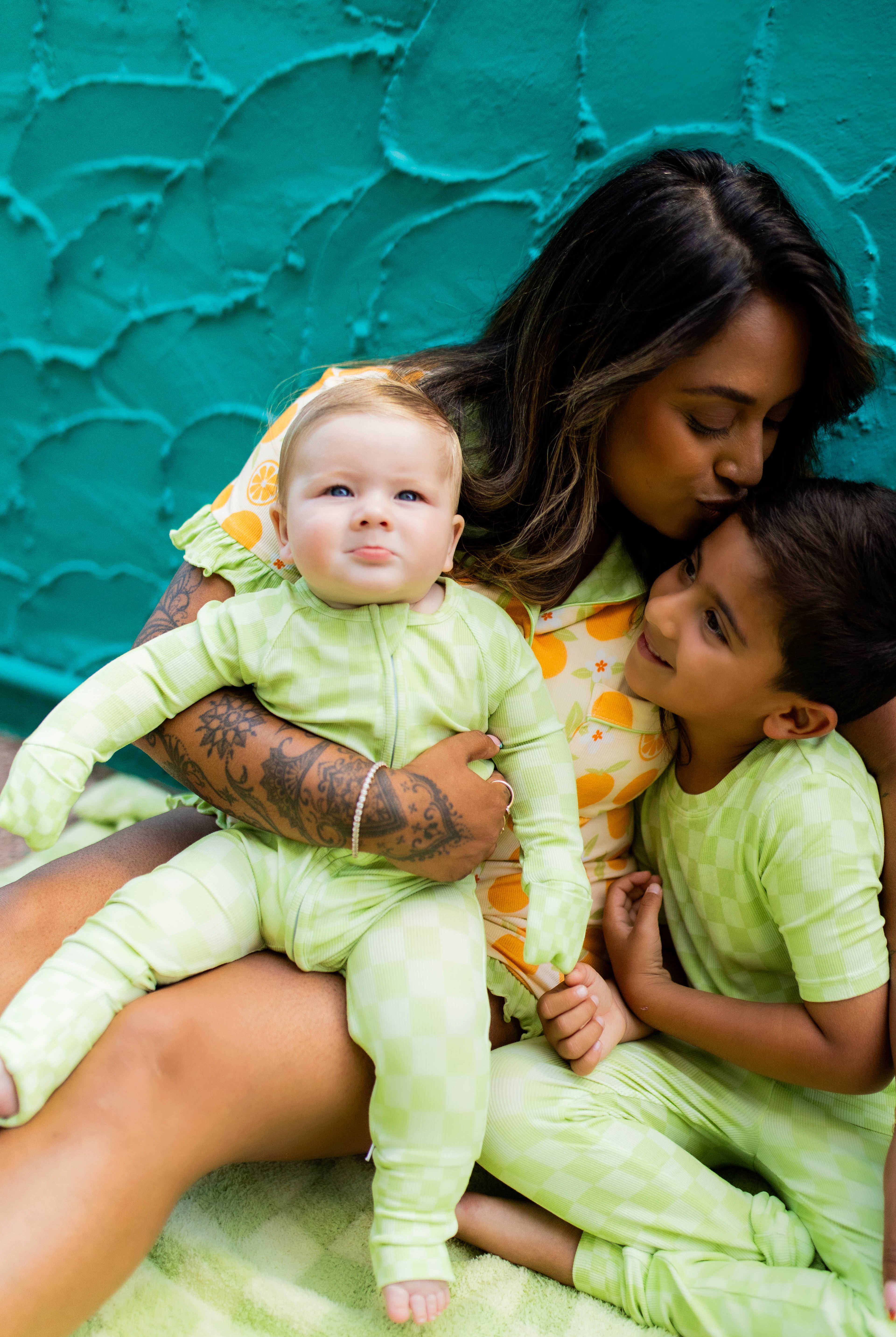 A mother and two children wearing matching light green patterned pajamas, sitting together against a textured teal wall.