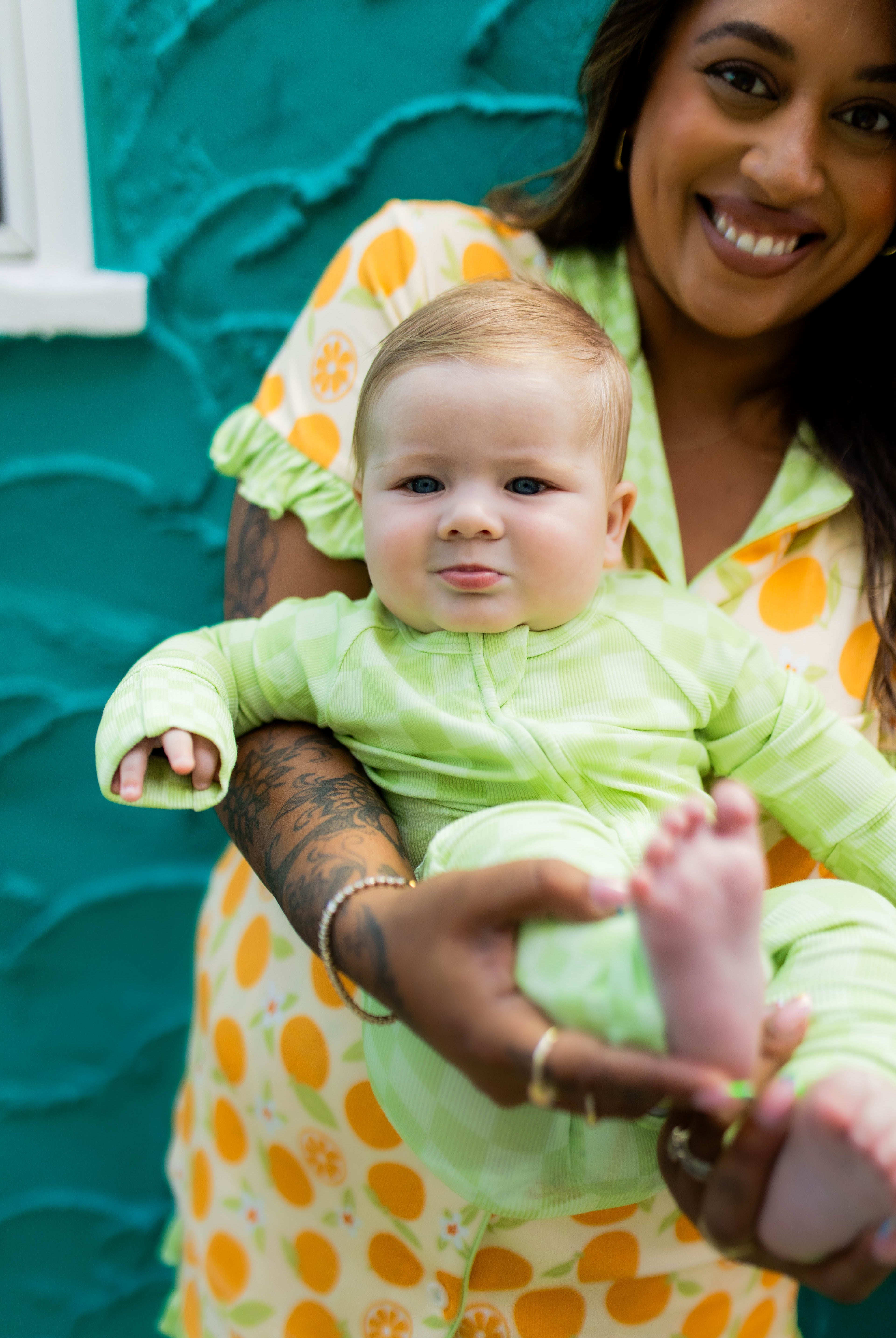 A smiling woman holds a baby wearing a light green long-sleeve outfit against a textured teal wall. The woman wears orange pjs.