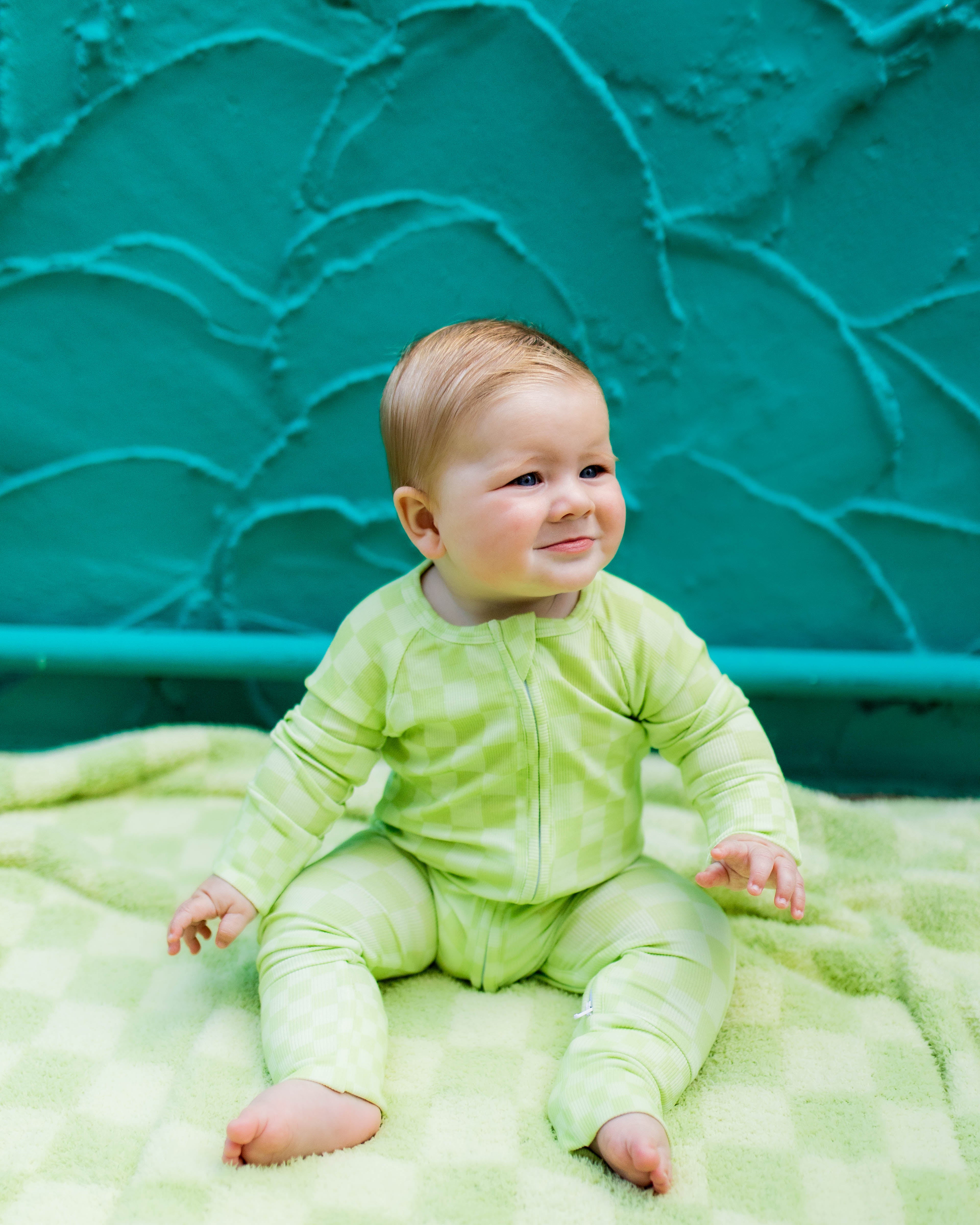 A baby wearing a light green long-sleeve romper sits on a green checkered blanket in front of a textured teal wall.