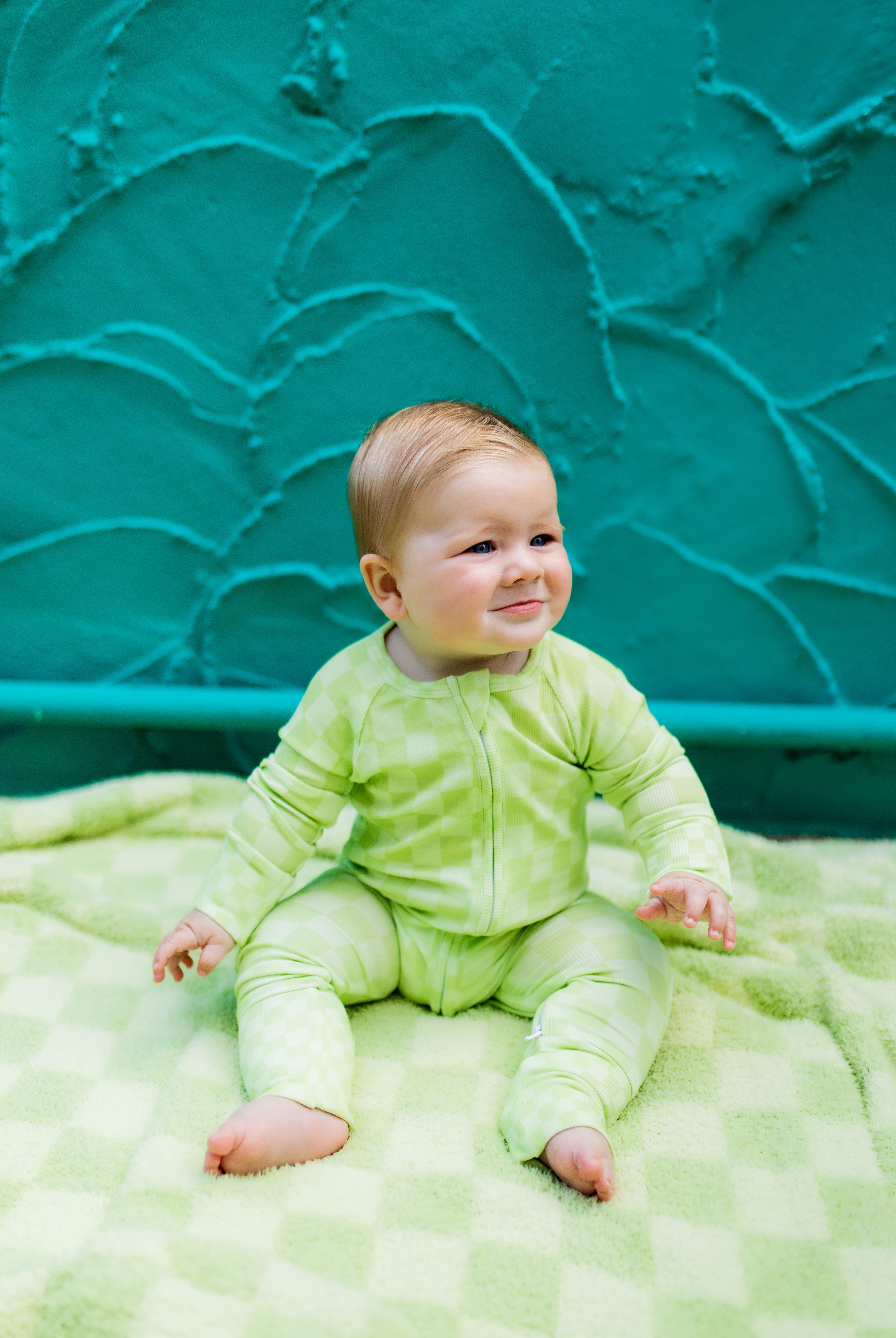 A baby wearing a light green long-sleeve romper sits on a green checkered blanket in front of a textured teal wall.
