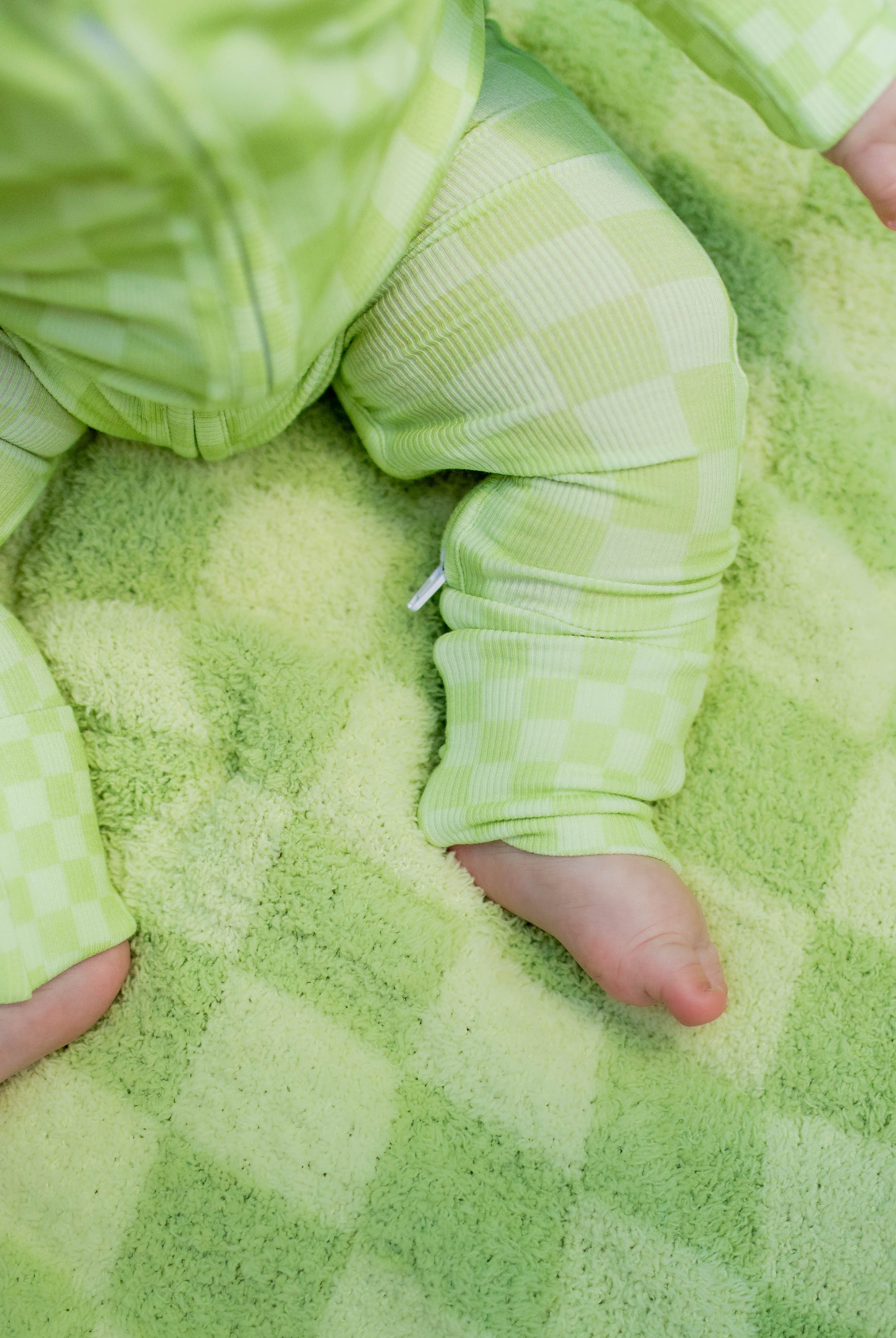 A baby wearing a light greencheckered one-piece outfit sits on a matching blanket.