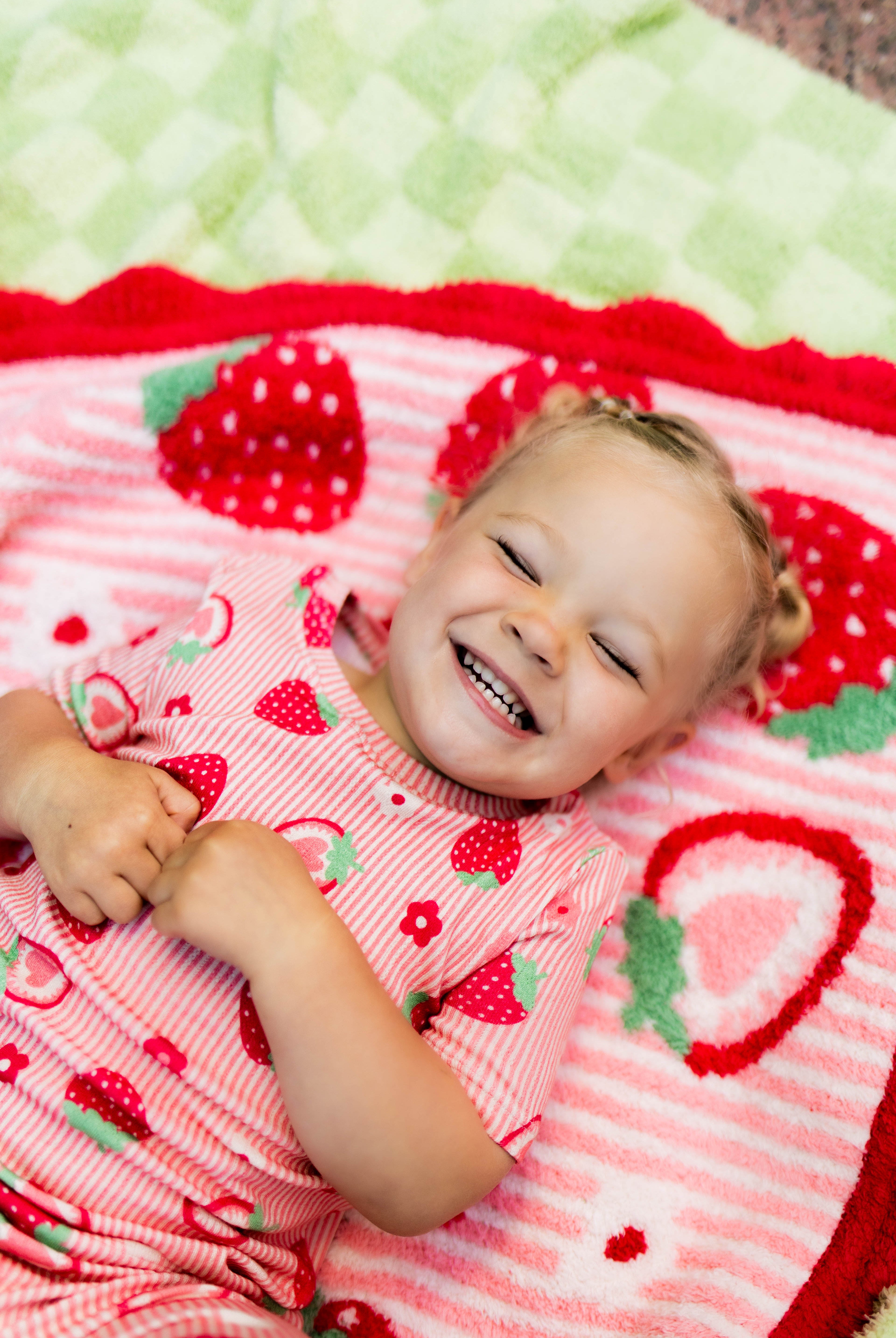 A smiling toddler lying on a strawberry-themed blanket, wearing a matching pink strawberry-print pajama top. The scene is styled with a cozy, playful aesthetic.