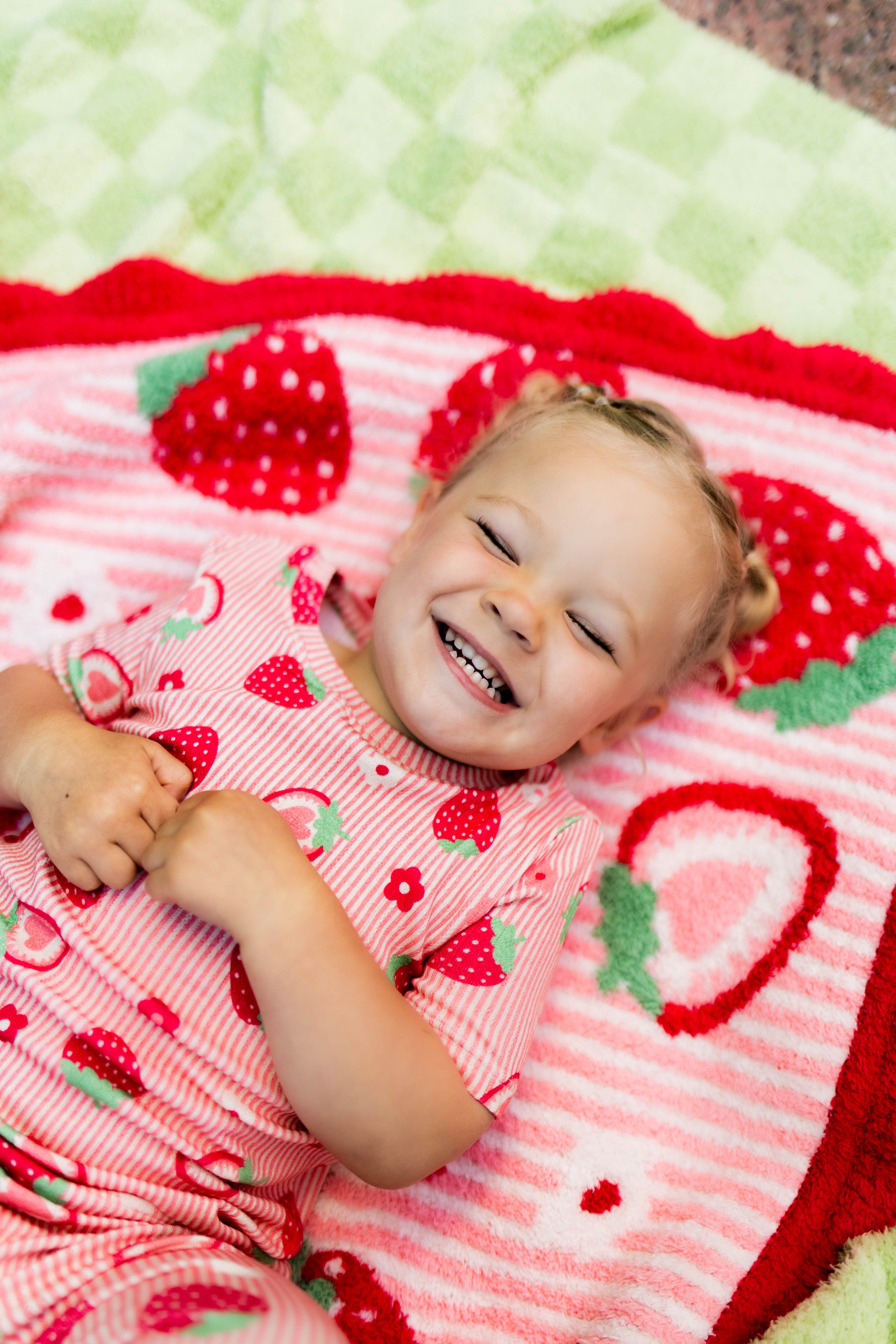 A smiling toddler lying on a strawberry-themed blanket, wearing a matching pink strawberry-print pajama top. The scene is styled with a cozy, playful aesthetic.