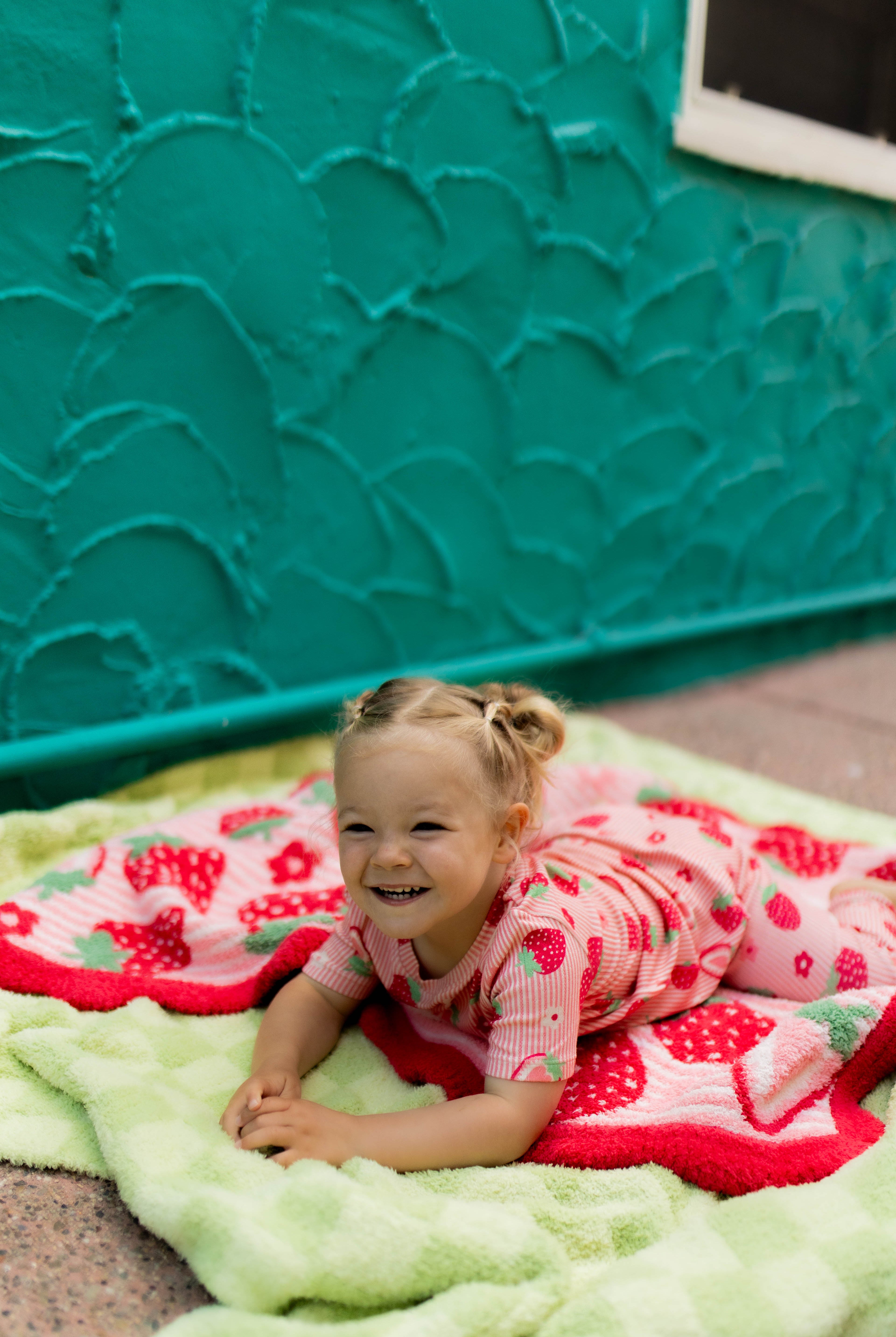 A smiling young girl lies on a light green plush blanket with a red and white strawberry-patterned blanket on top of it posed in front of a textured teal wall.