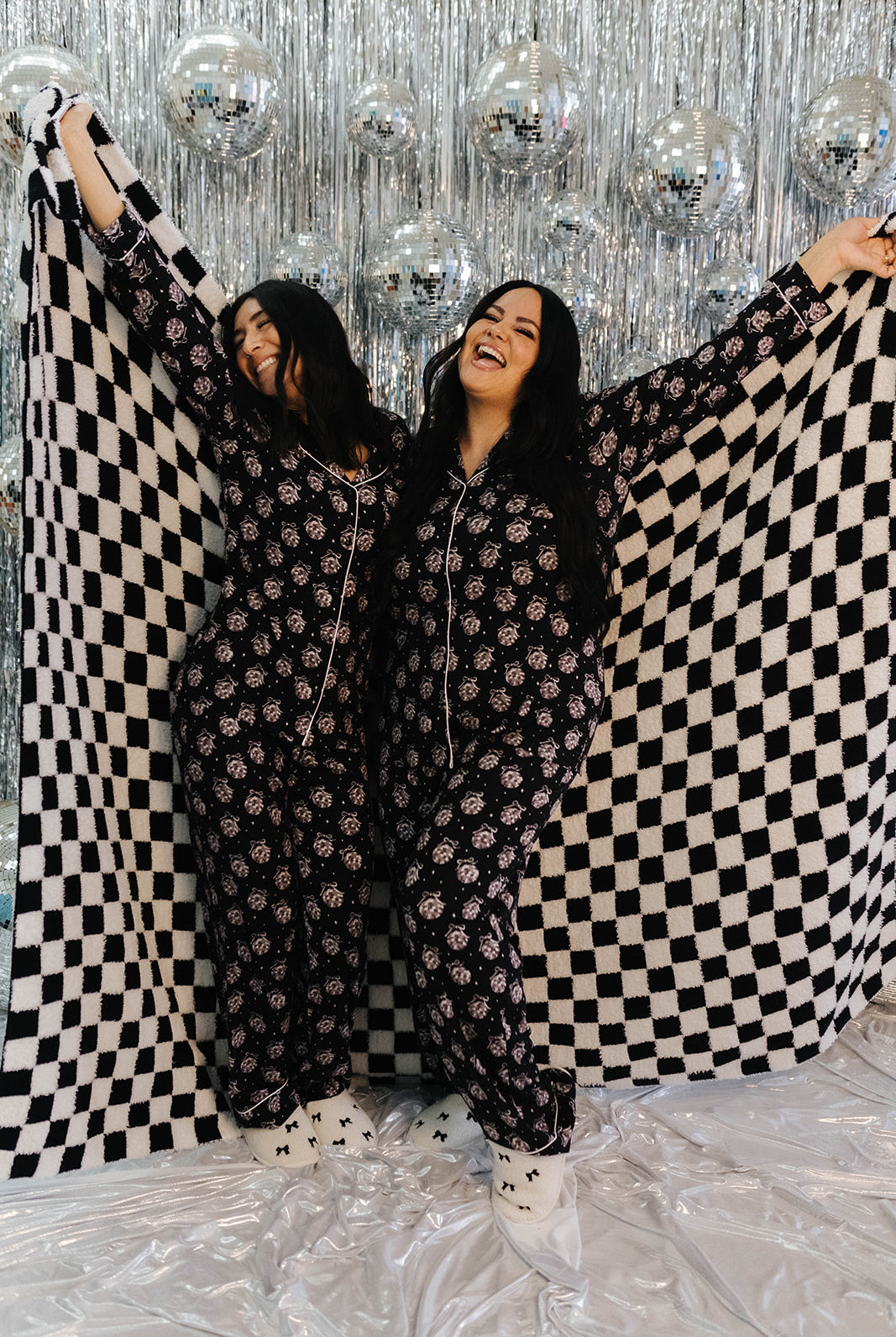 Two women holding a black and white checkered blanket against a silver disco ball background